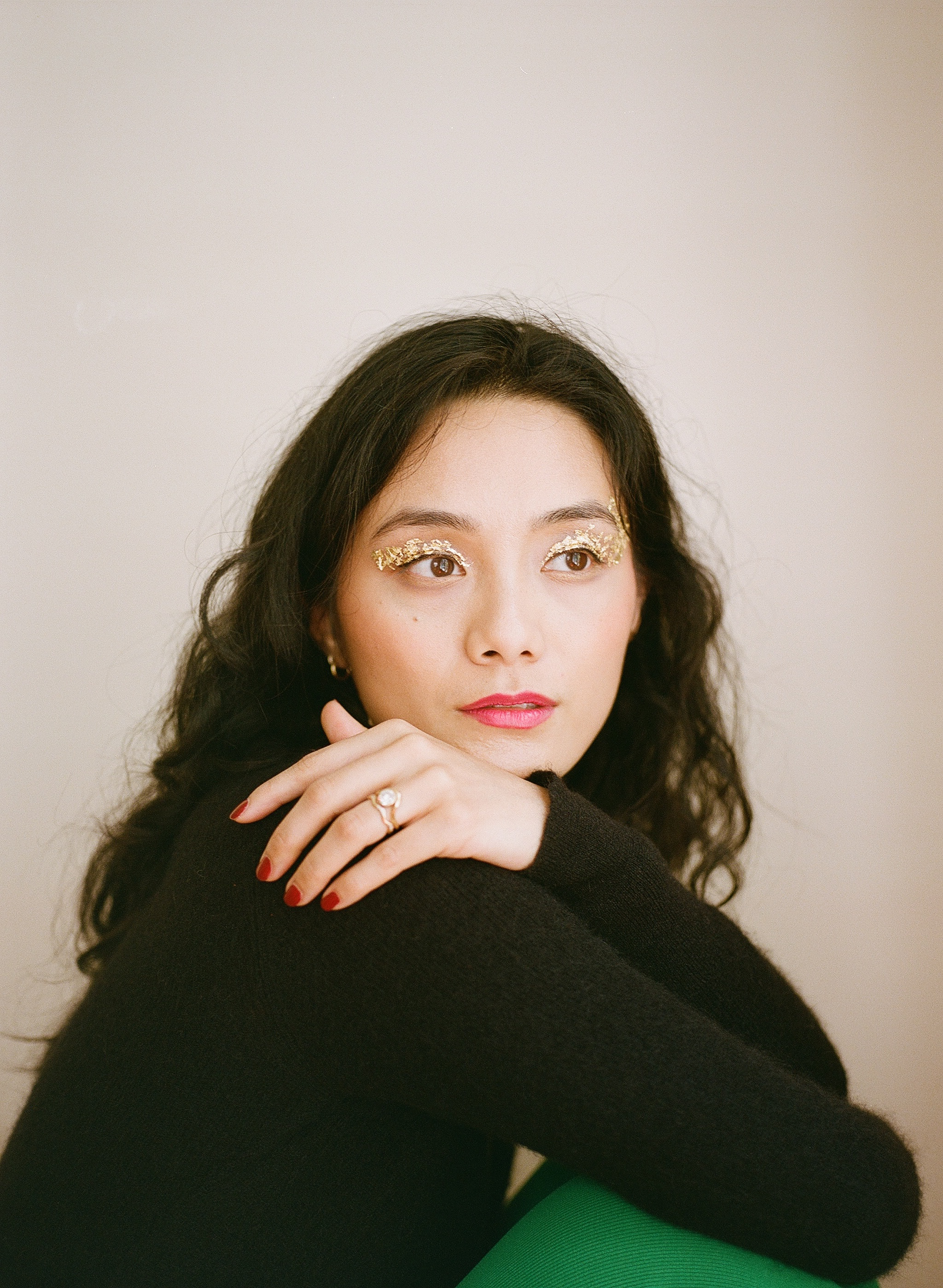 Portrait of a woman with long dark hair, glittery gold eye makeup, red lipstick, and red nails wearing a black sweater