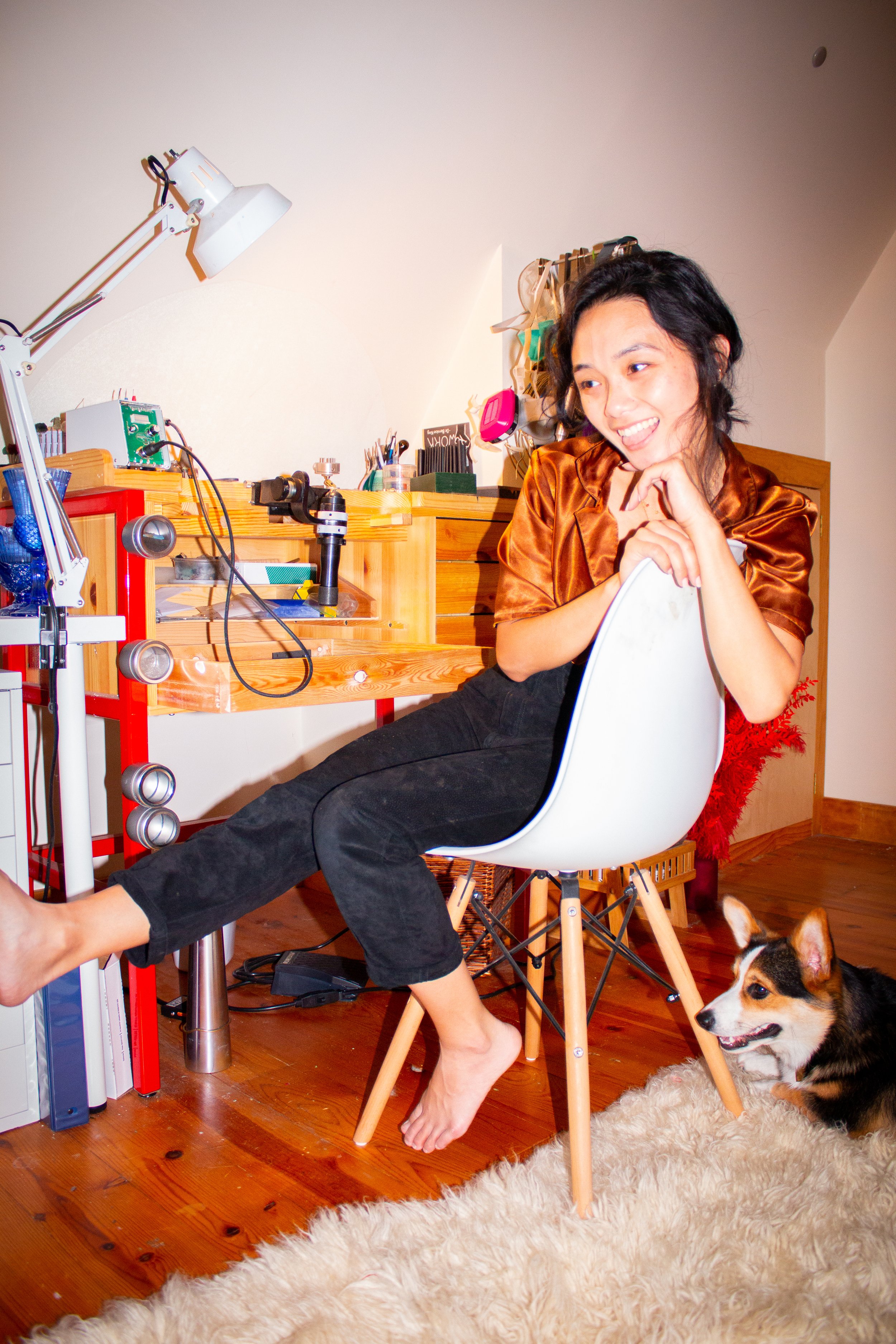 Smiling person in a home workshop beside a workbench, seated in a white chair with a dog on a rug nearby
