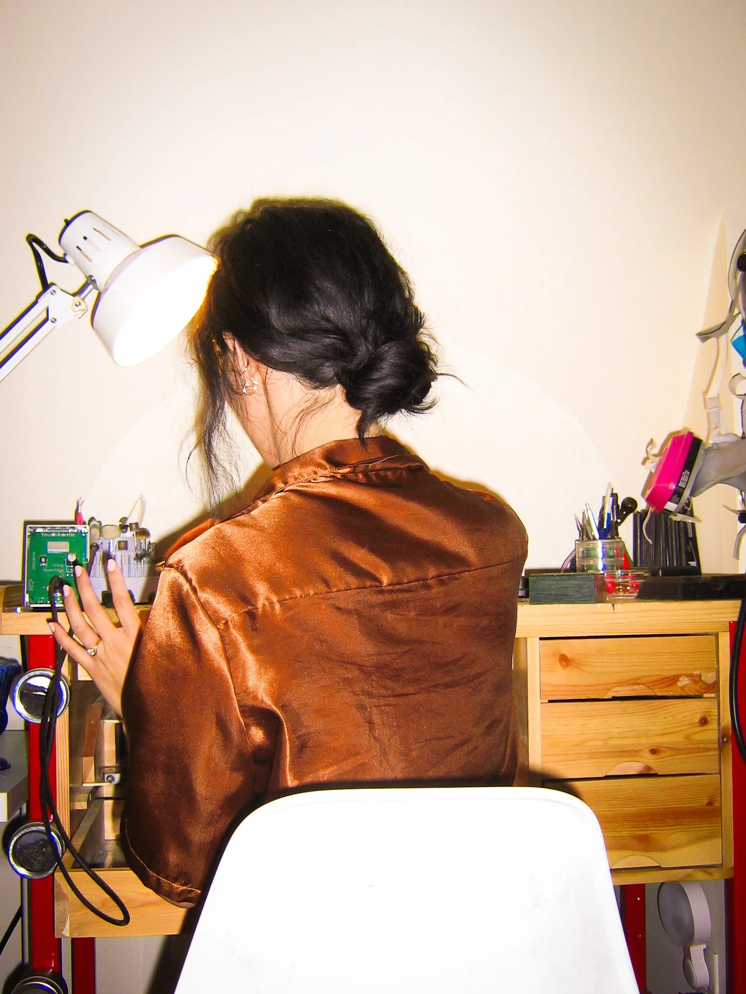 Woman in a brown satin jacket working at a desk with electronic equipment, tools, and a lamp