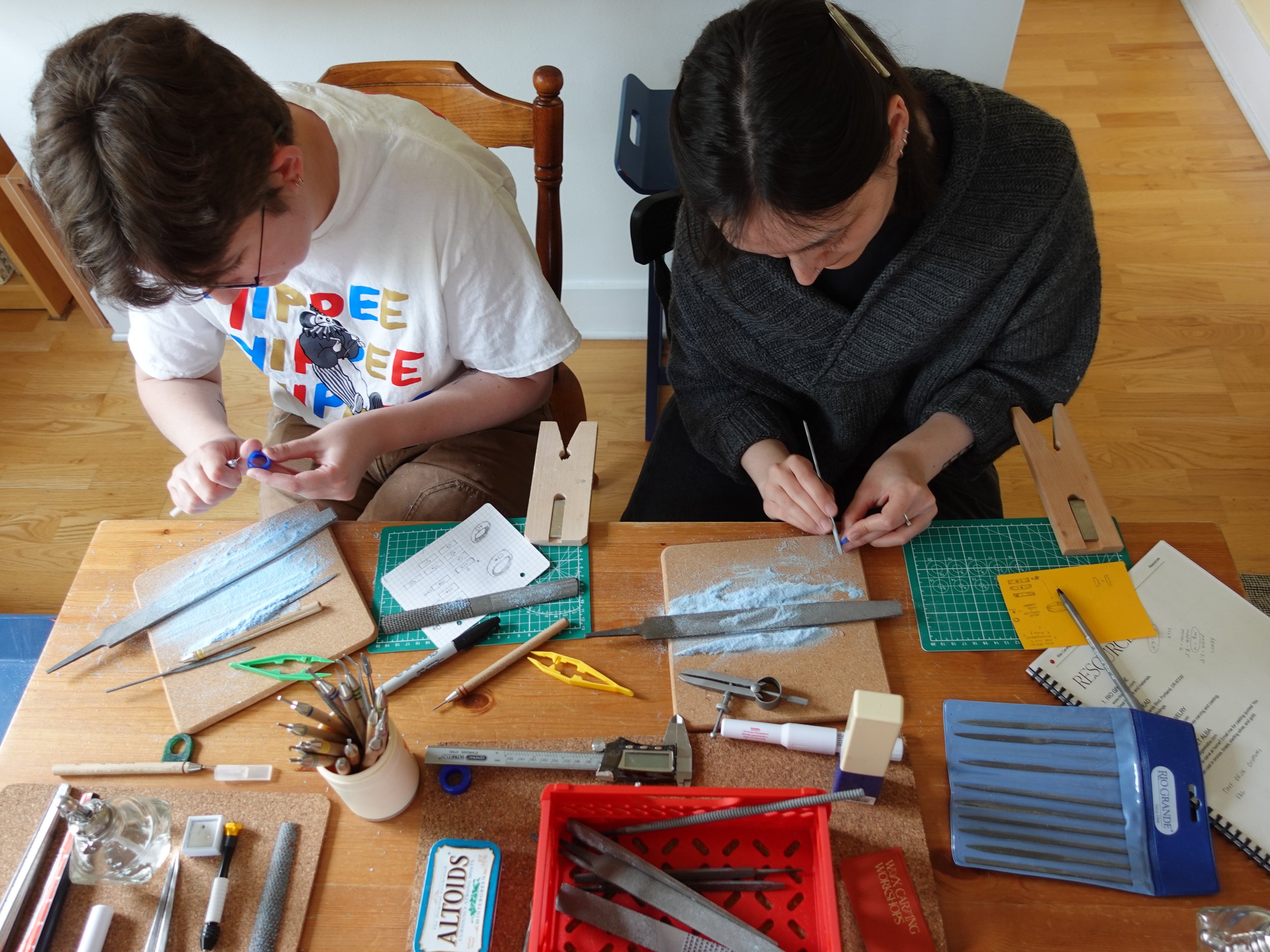 Two people work on a jewelry-making project at a table covered with files, carving tools, cutting mats, and other craft supplies.