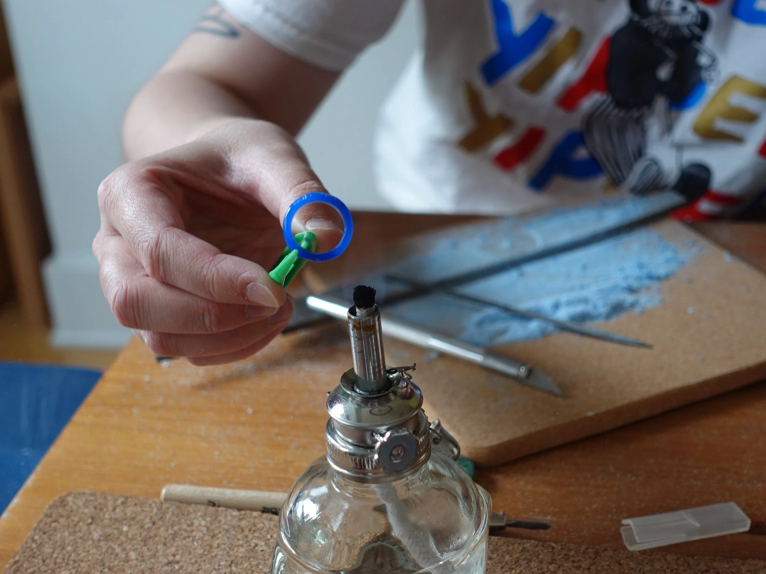 Close-up of a person using a green-handled tool over an alcohol lamp on a craft workbench