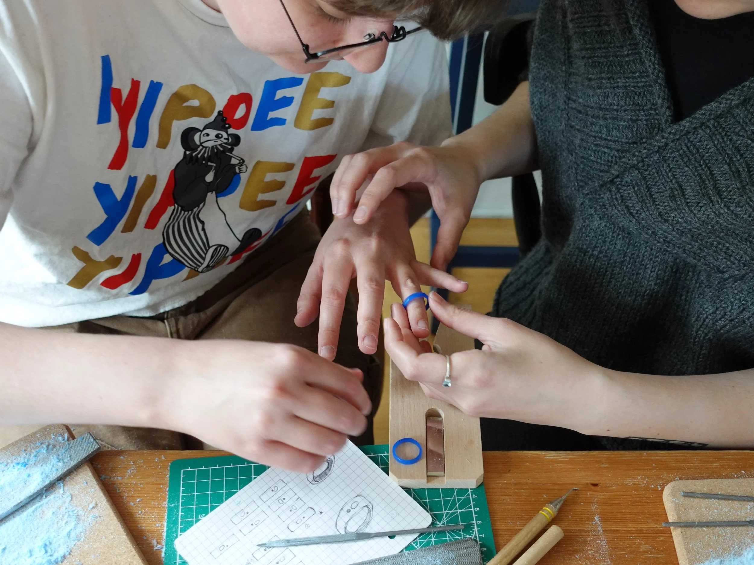 People crafting a blue ring at a workbench with tools, sandpaper, and design sketches