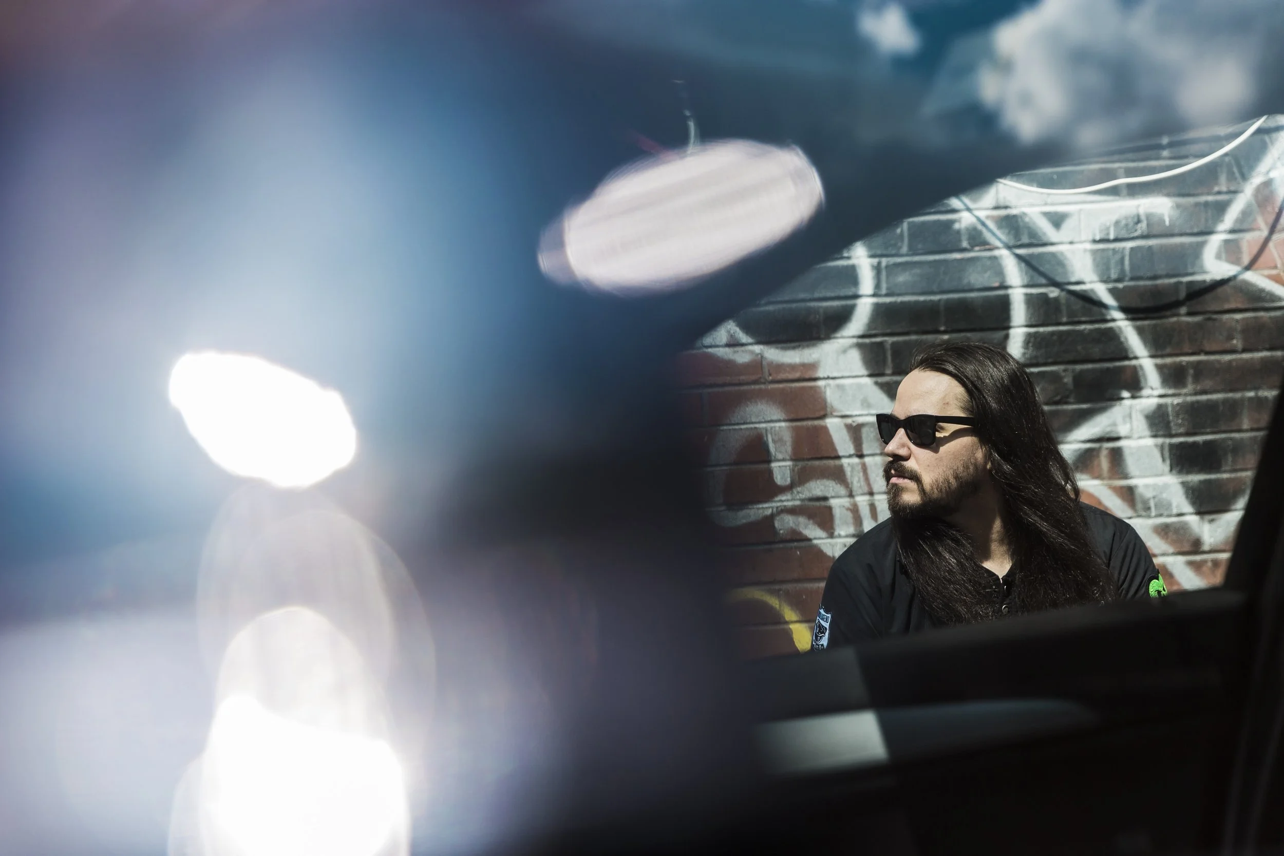Long-haired man wearing sunglasses and a black shirt standing in front of a graffiti-covered brick wall