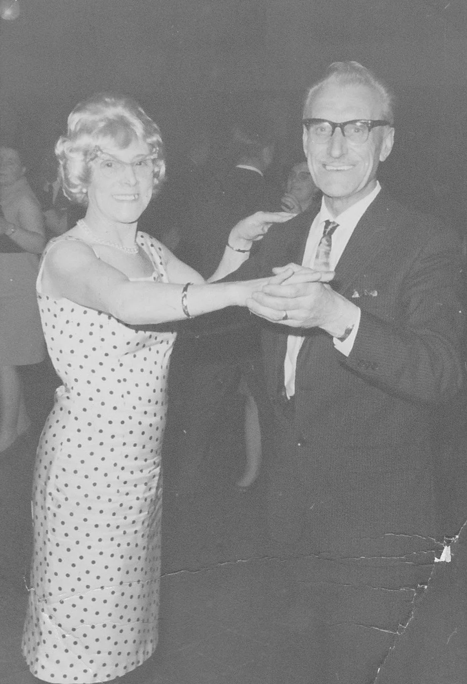 An old black and white photo of an elderly woman and a man dancing together, holding hands. The woman is wearing a polka dot dress and the man is dressed in a suit with glasses. They are smiling and appear to be in a social gathering or dance event.