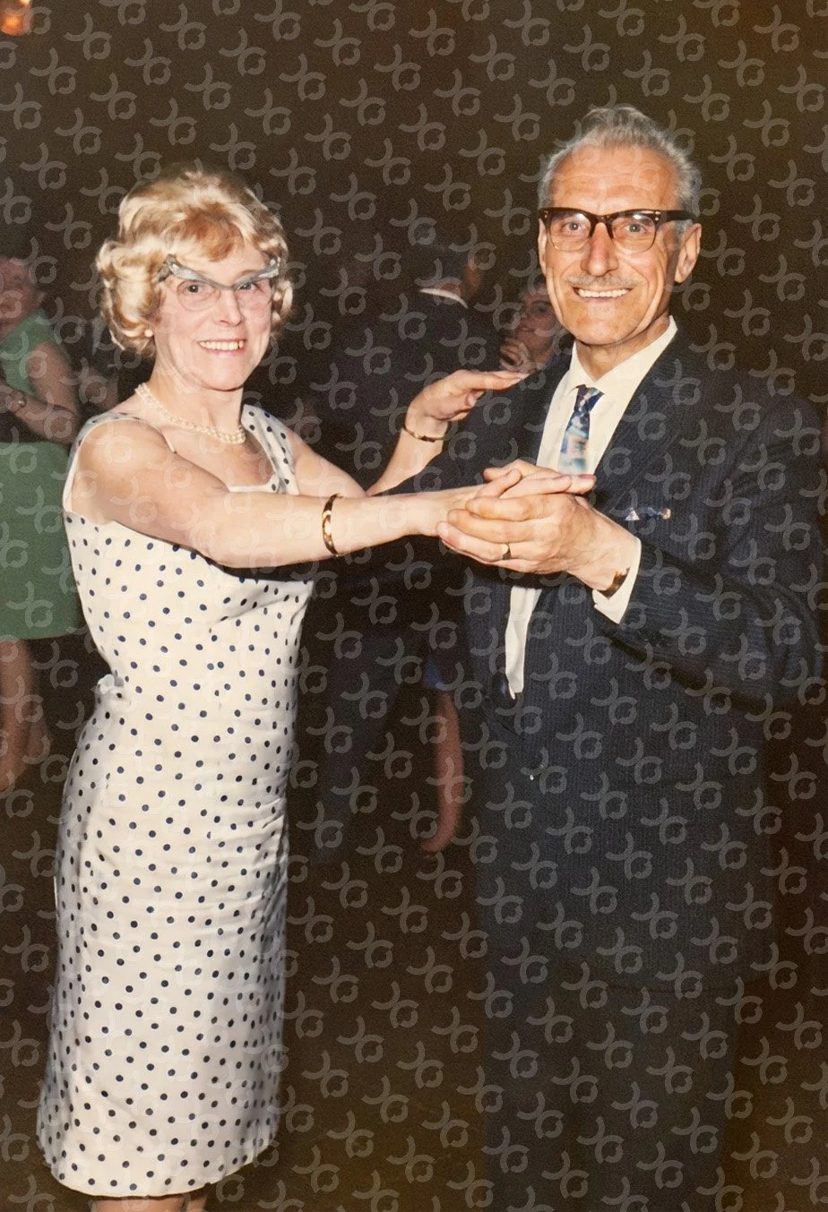 A restored colour photo of an elderly woman and a man dancing together, holding hands. The woman is wearing a polka dot dress and the man is dressed in a suit with glasses. They are smiling and appear to be in a social gathering or dance event.