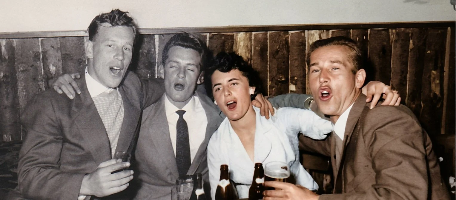 Four young people in vintage attire, sitting closely together in a bar or pub, singing or shouting while holding bottles and glasses of beer, with a wooden wall in the background. Half of the image is black and white and half is colour.