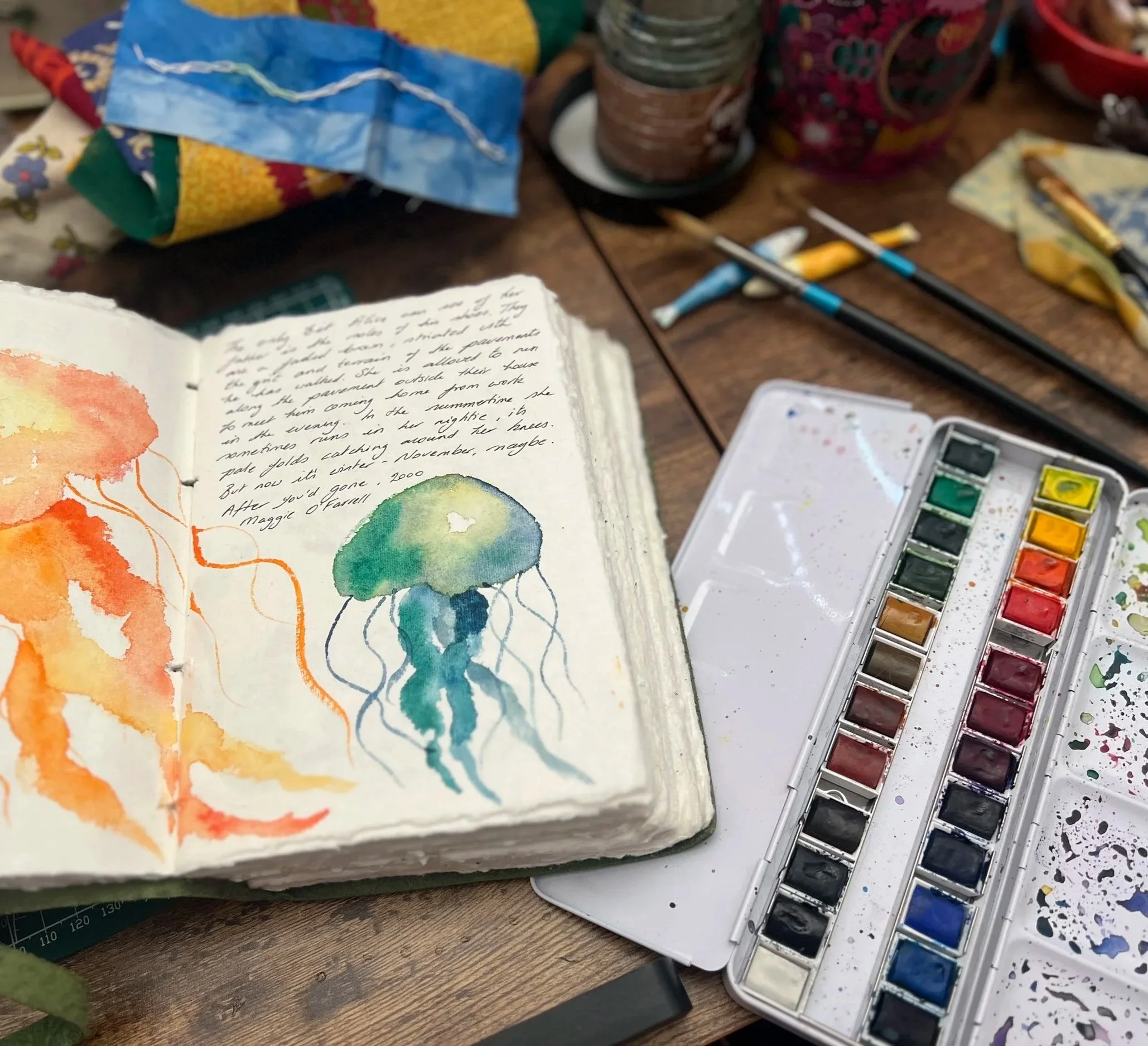 Open watercolor journal with handwritten notes and floral painting beside watercolor palettes and brushes on a wooden art table