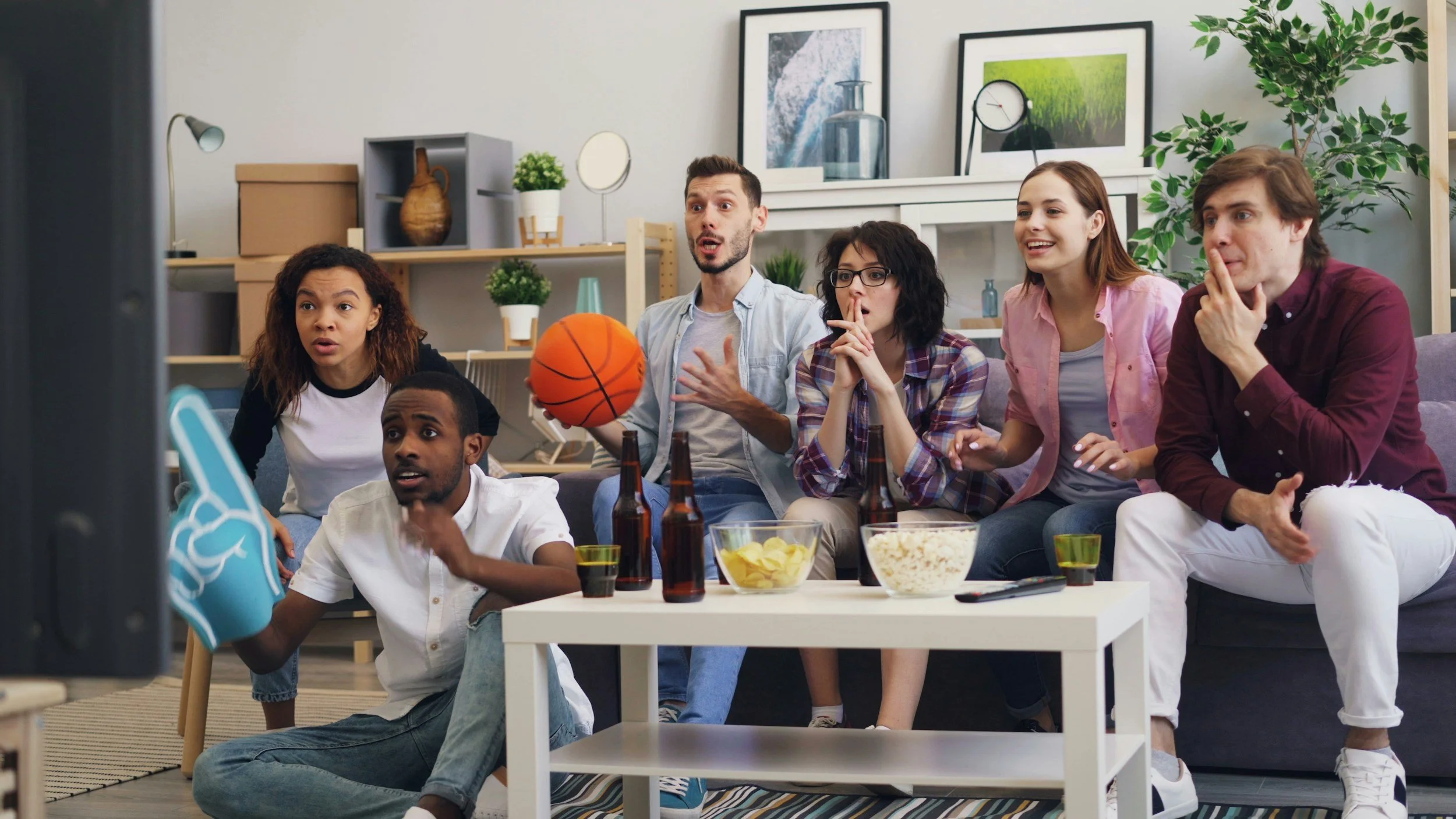 Diverse group of friends watching a basketball game together in a living room with snacks and drinks.
