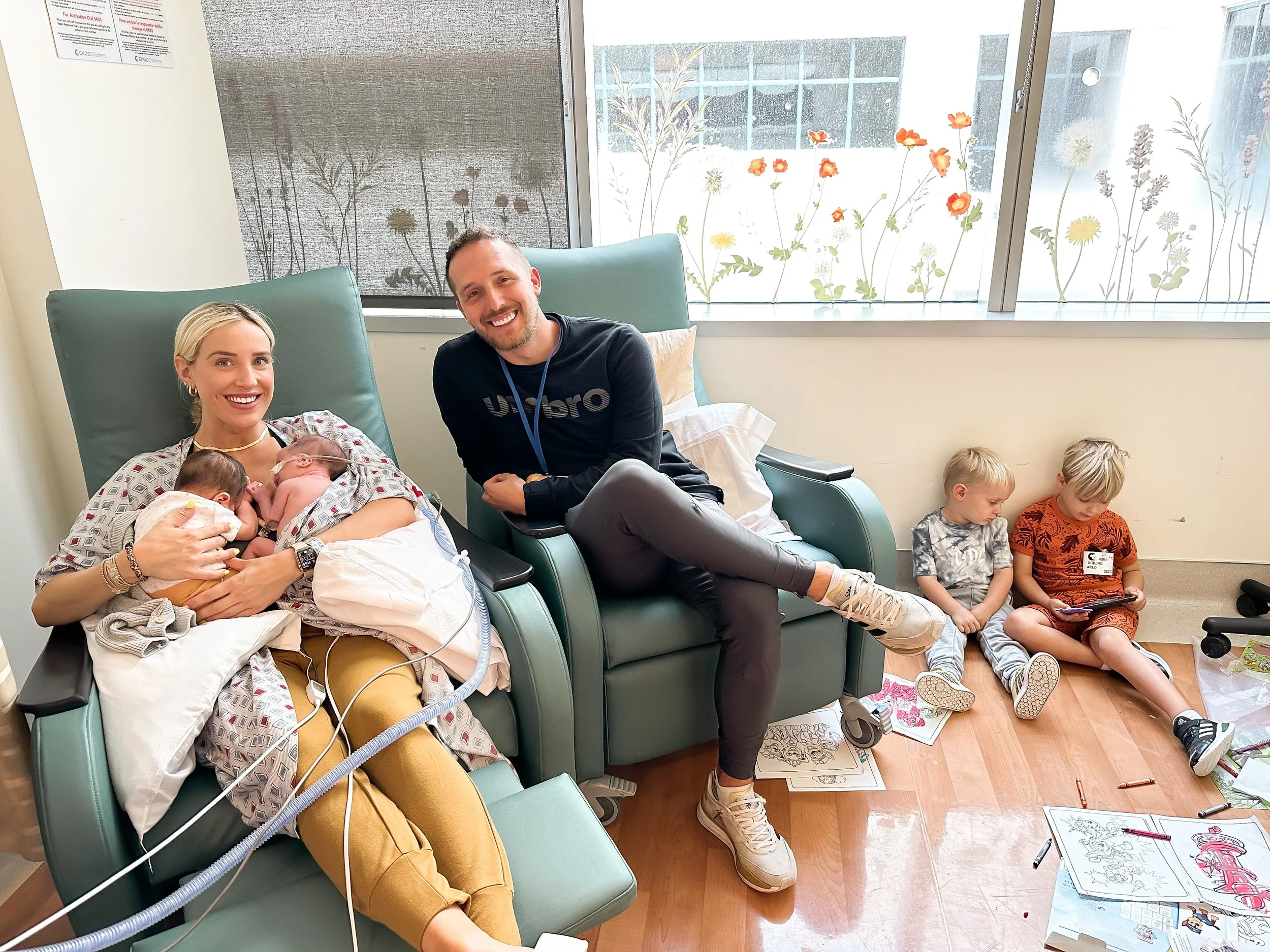 Family gathered in a hospital room, with a mother holding newborn twins, a seated father, and two young children coloring nearby.