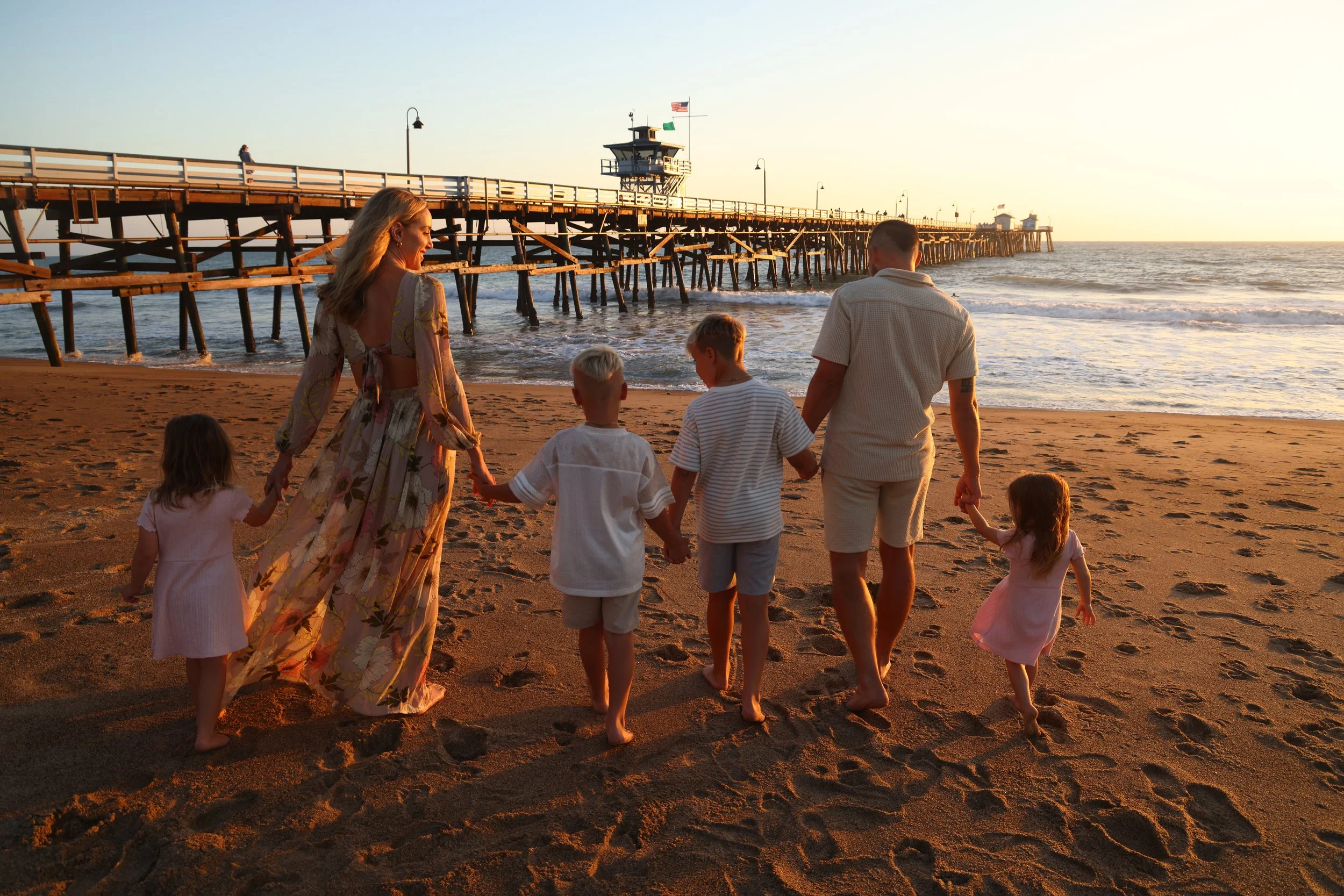 Family walking on a sandy beach at sunset beside a wooden ocean pier with lifeguard tower and flags