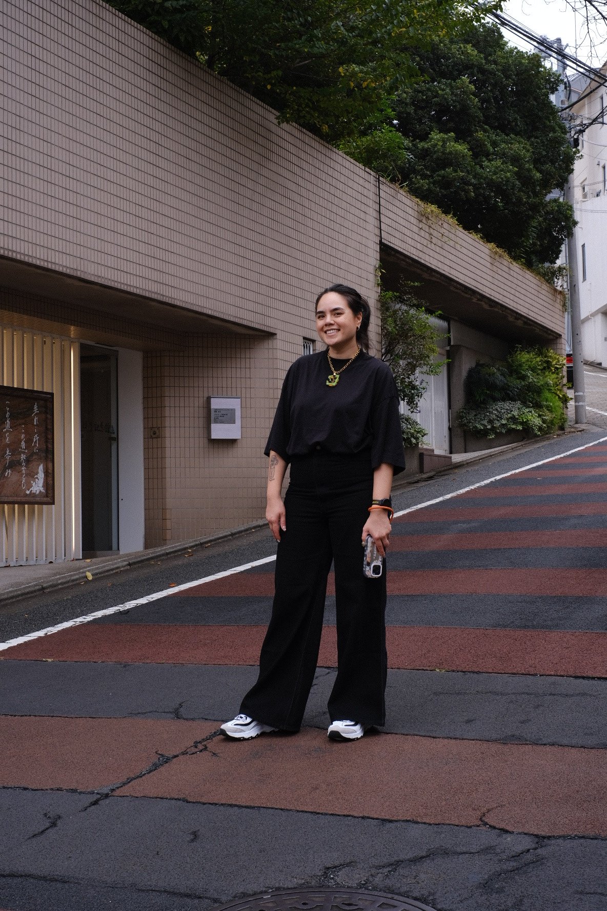 Woman in black clothing and white sneakers standing on a city street beside a tiled building