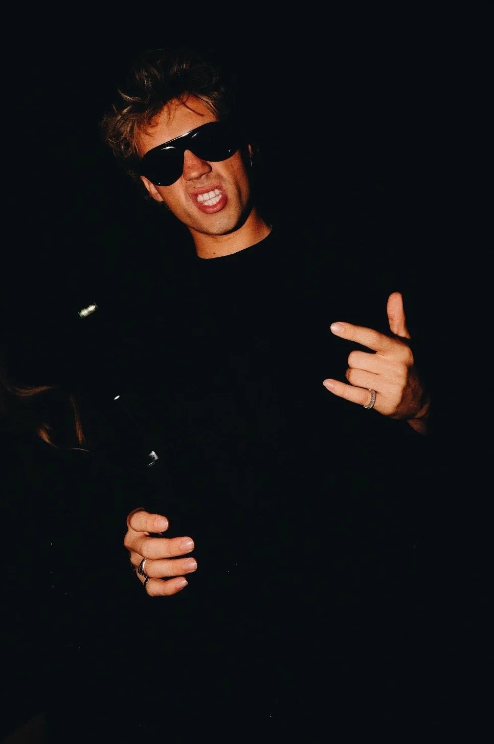 Person wearing black sunglasses and clothing, making hand gestures against a dark background