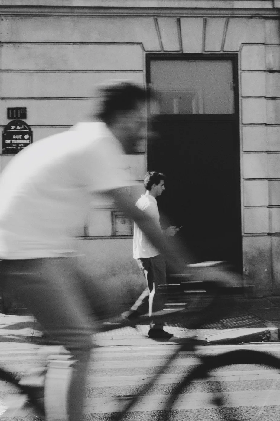 Black-and-white street photograph of blurred pedestrians walking past a doorway on a city sidewalk.