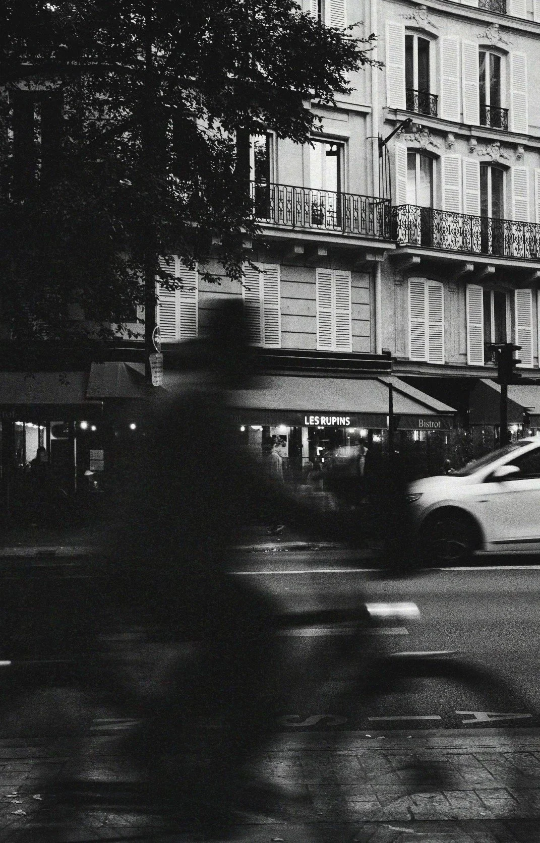 Black-and-white street scene showing a Parisian building with ornate balconies, the Les Rupins bistro, a passing car, and a tree in the foreground.