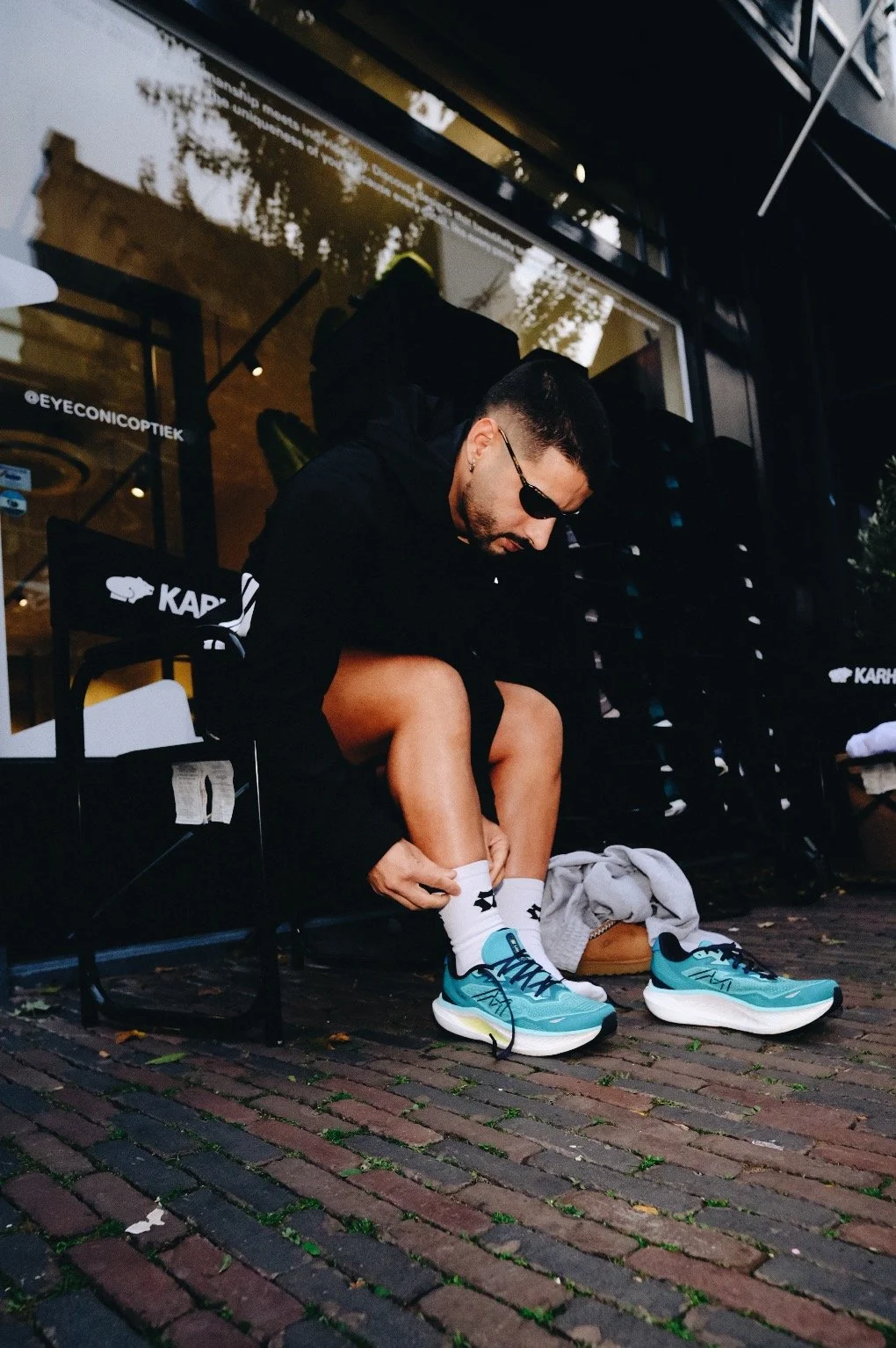 Man wearing sunglasses adjusts his white socks beside turquoise running shoes outside an eyewear store.