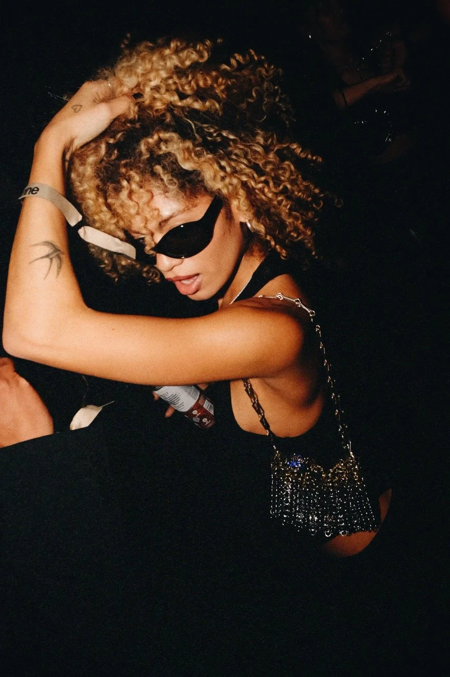 Woman with curly hair wearing black sunglasses and a chain purse in a dimly lit setting
