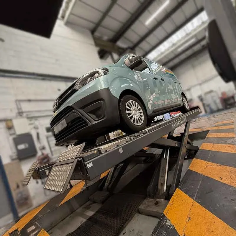 Light blue compact car undergoing testing on a raised dynamometer platform inside an automotive workshop