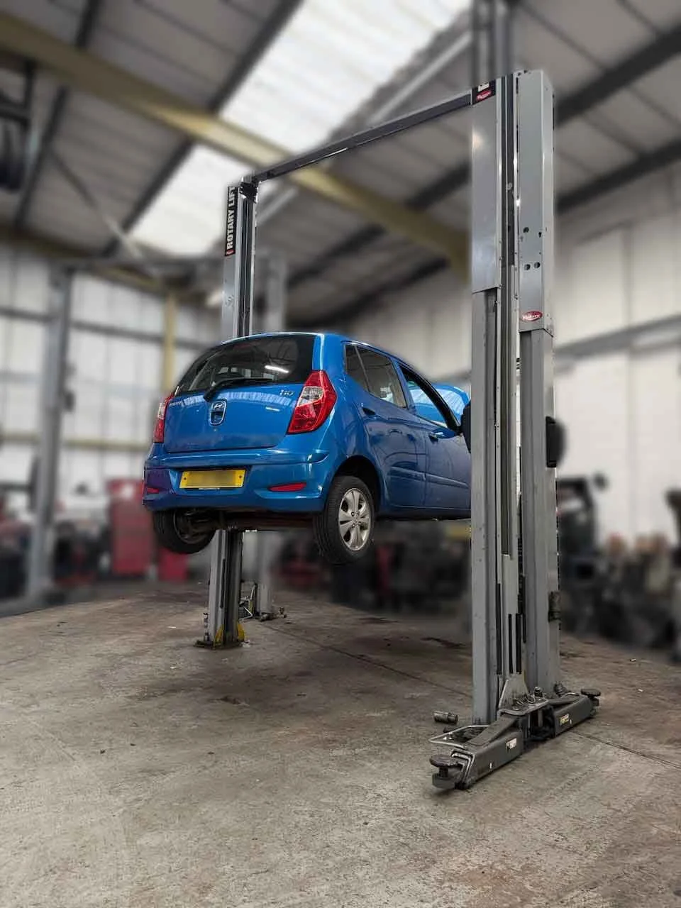 Blue Hyundai i10 raised on a two-post vehicle lift in an auto repair shop