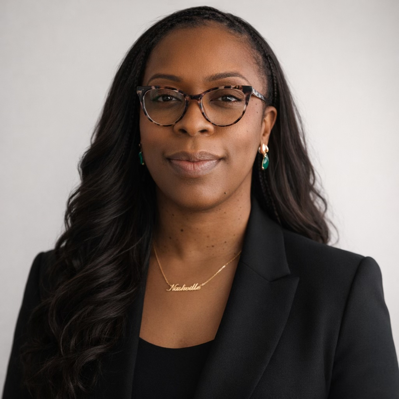 Professional headshot of a Black woman wearing glasses, a black blazer, green drop earrings, and a nameplate necklace
