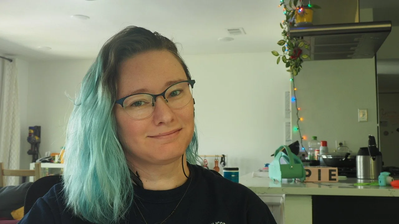 Smiling person with teal hair and glasses posing in a kitchen decorated with colorful string lights