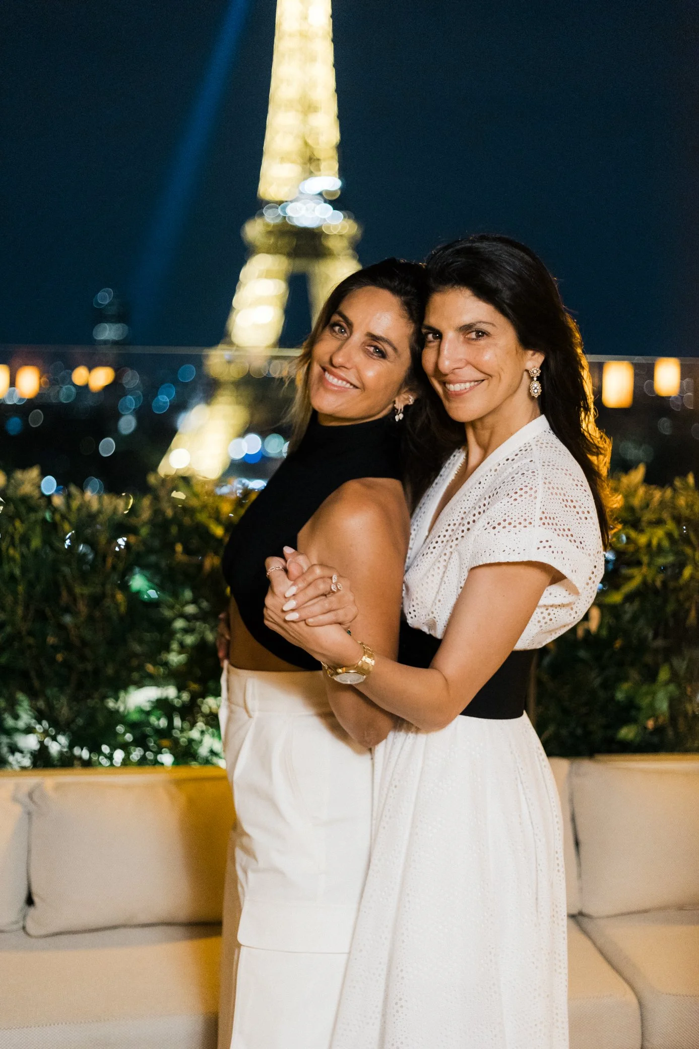 Deux femmes souriantes posant enlacées devant la tour Eiffel illuminée, de nuit.