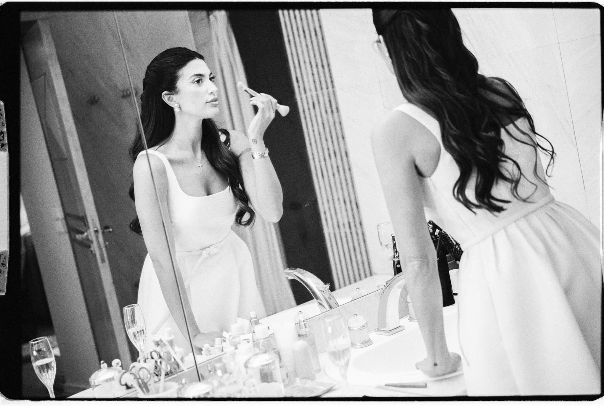 Femme en tenue blanche se maquillant devant un miroir de salle de bains, entourée de produits de beauté.