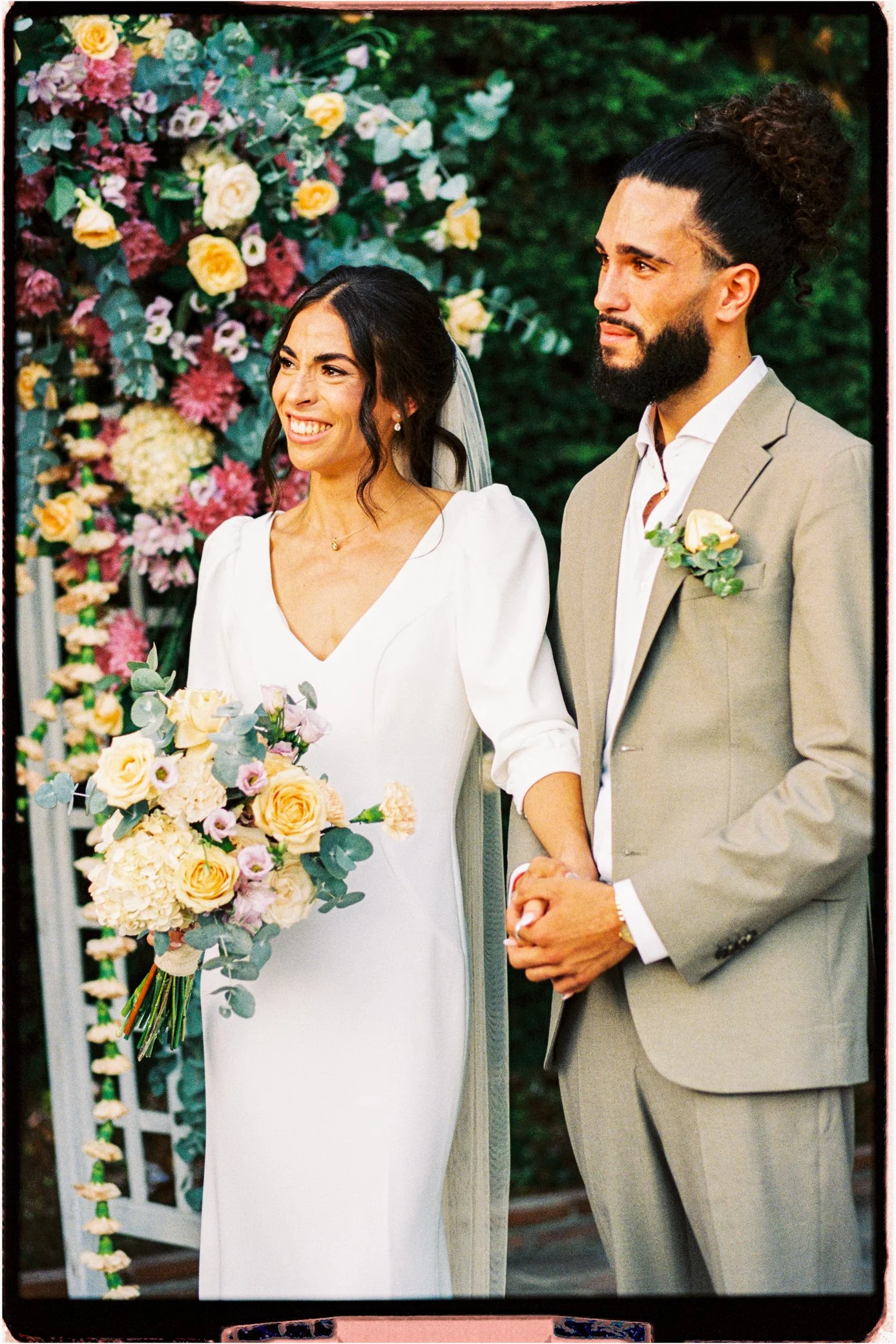 Couple de mariés souriants devant une arche fleurie, la mariée tenant un bouquet de roses et d'hortensias.