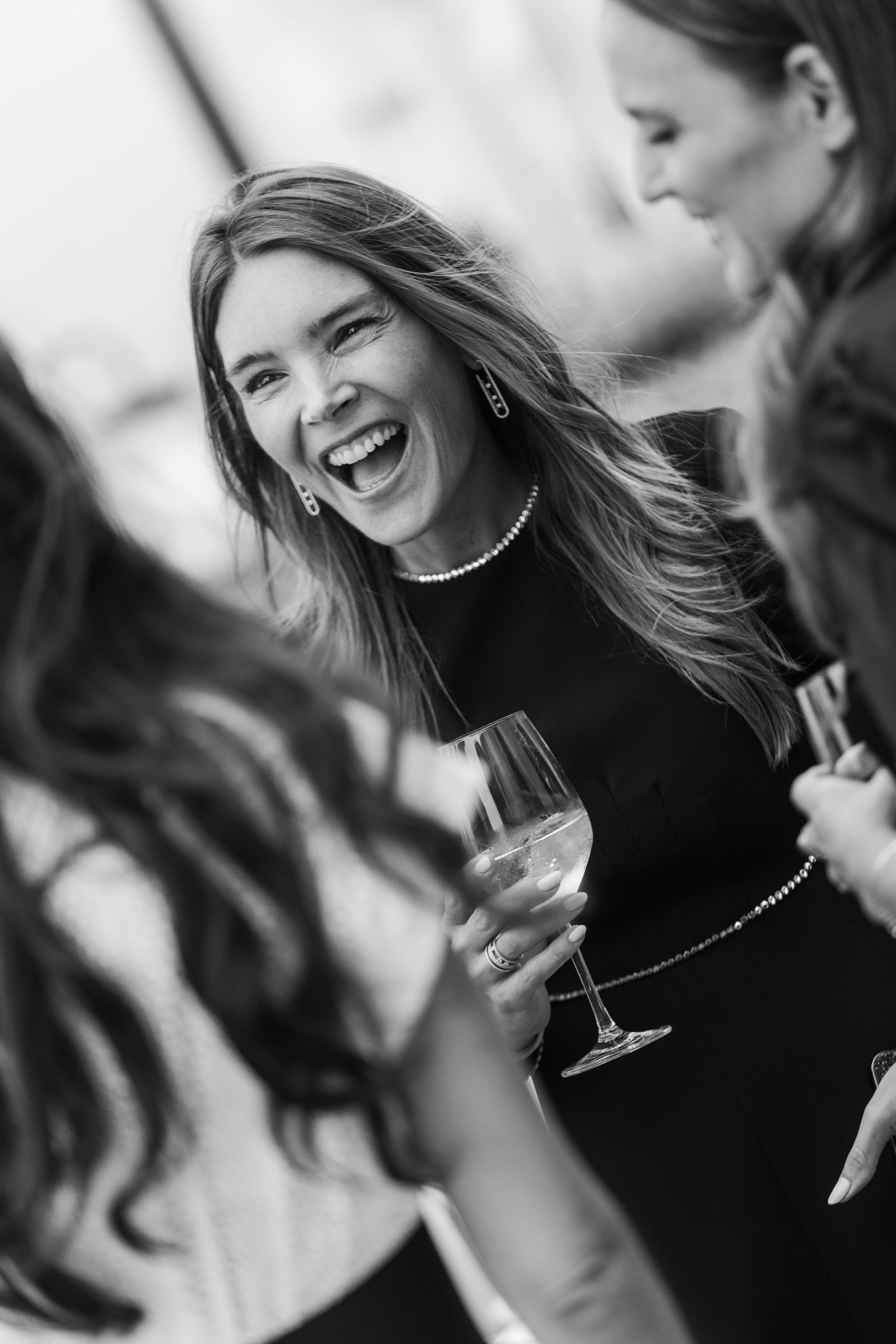 Femme souriante tenant un verre de vin lors d’une réception, entourée d’invités, en noir et blanc.