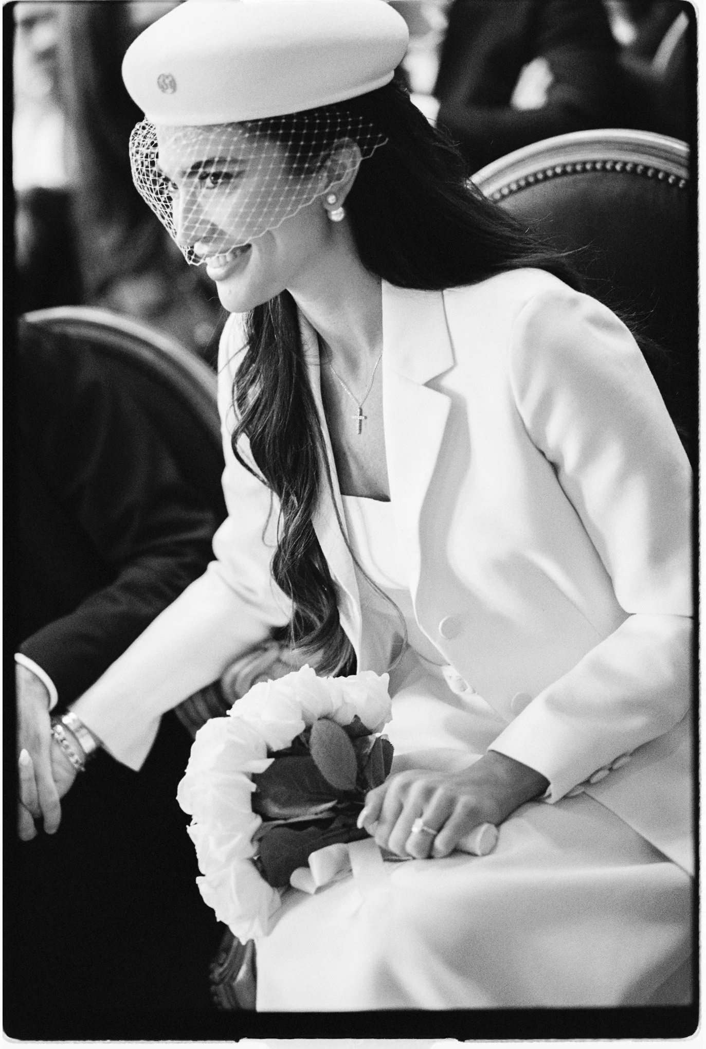 Femme portant un tailleur blanc, un chapeau à voile et tenant un bouquet de fleurs lors d’une cérémonie.