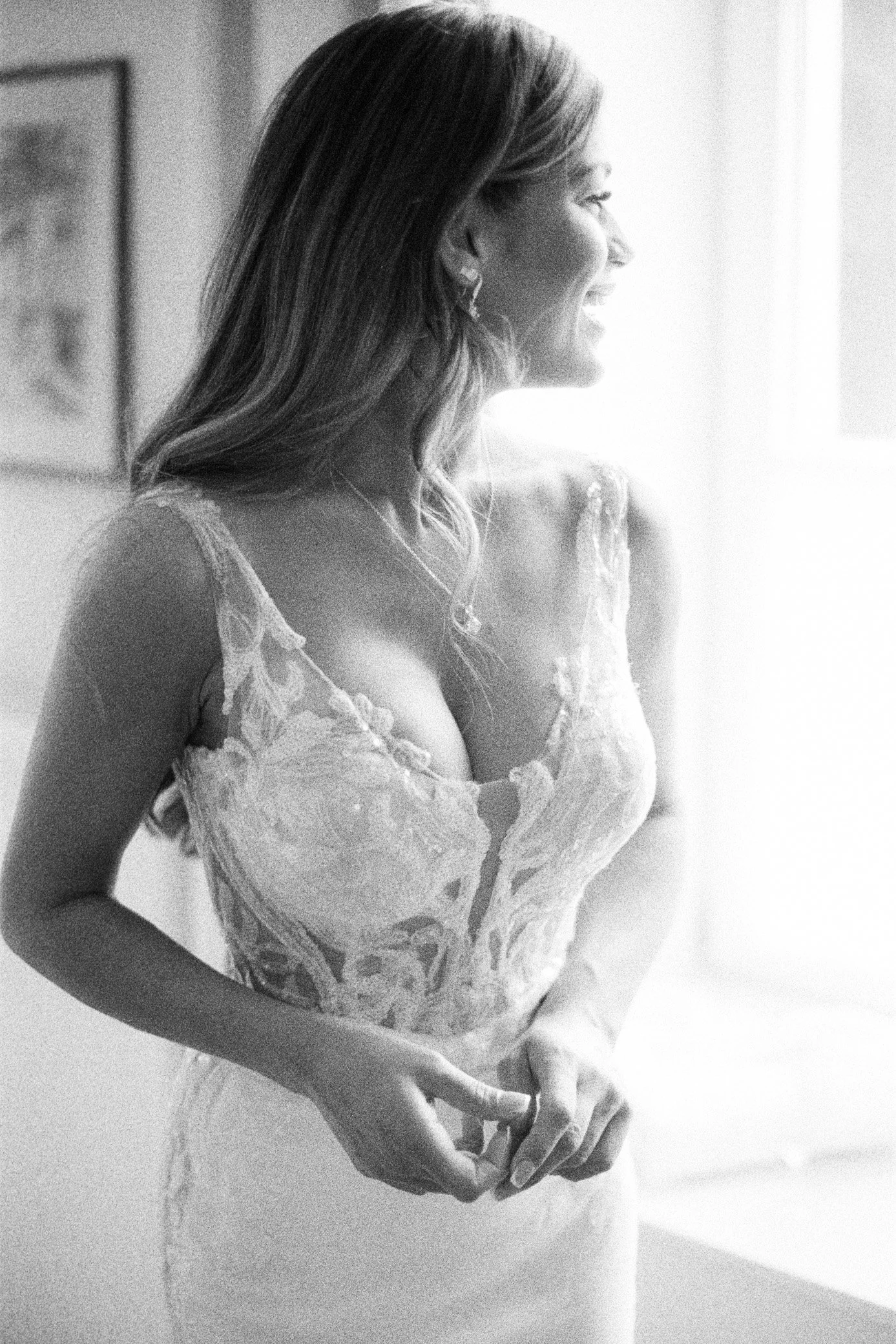 Femme en robe de mariée en dentelle, photographiée de profil près d’une fenêtre lumineuse.