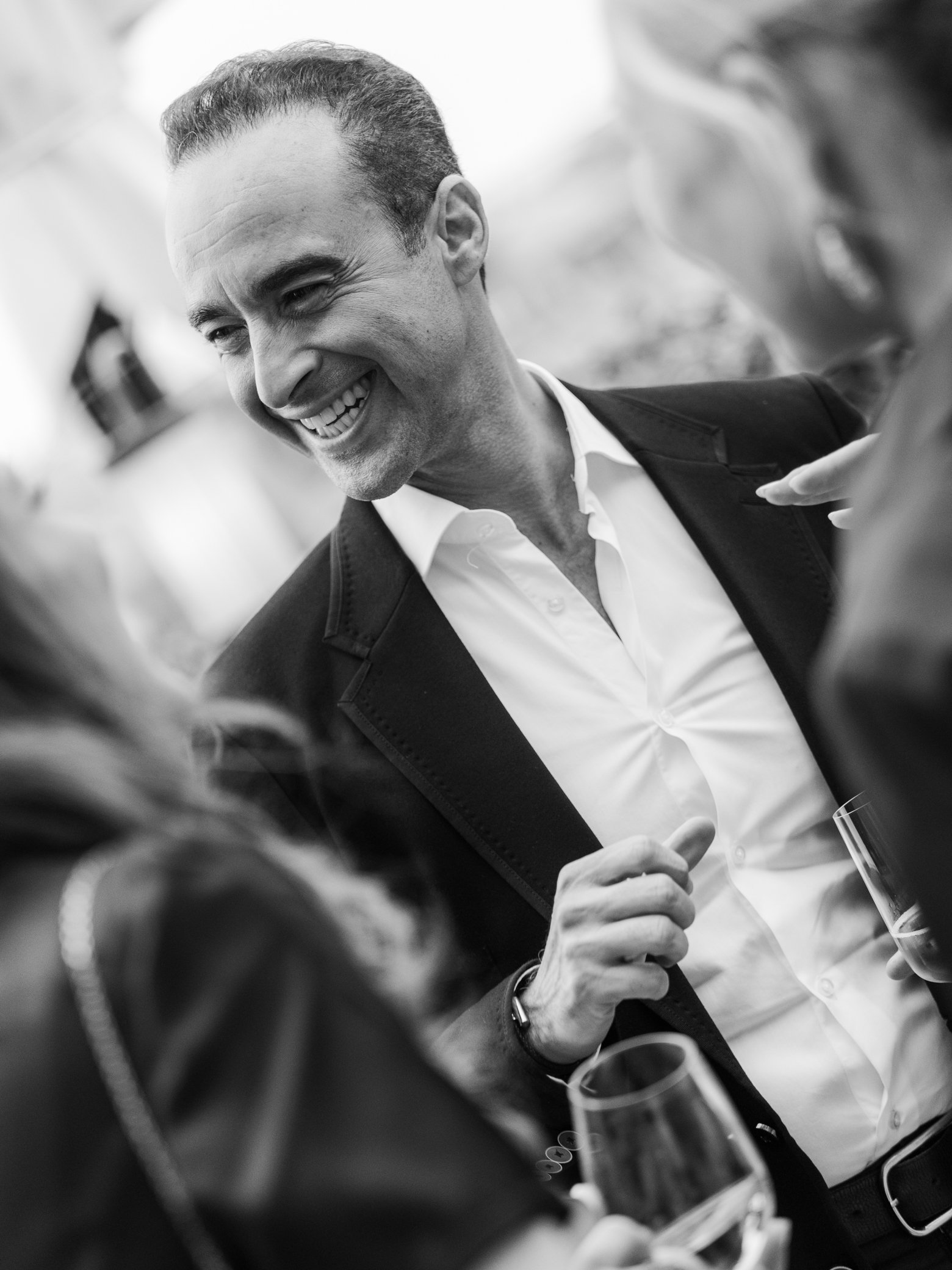 Homme souriant en costume lors d’une réception, tenant un verre au milieu d’invités.