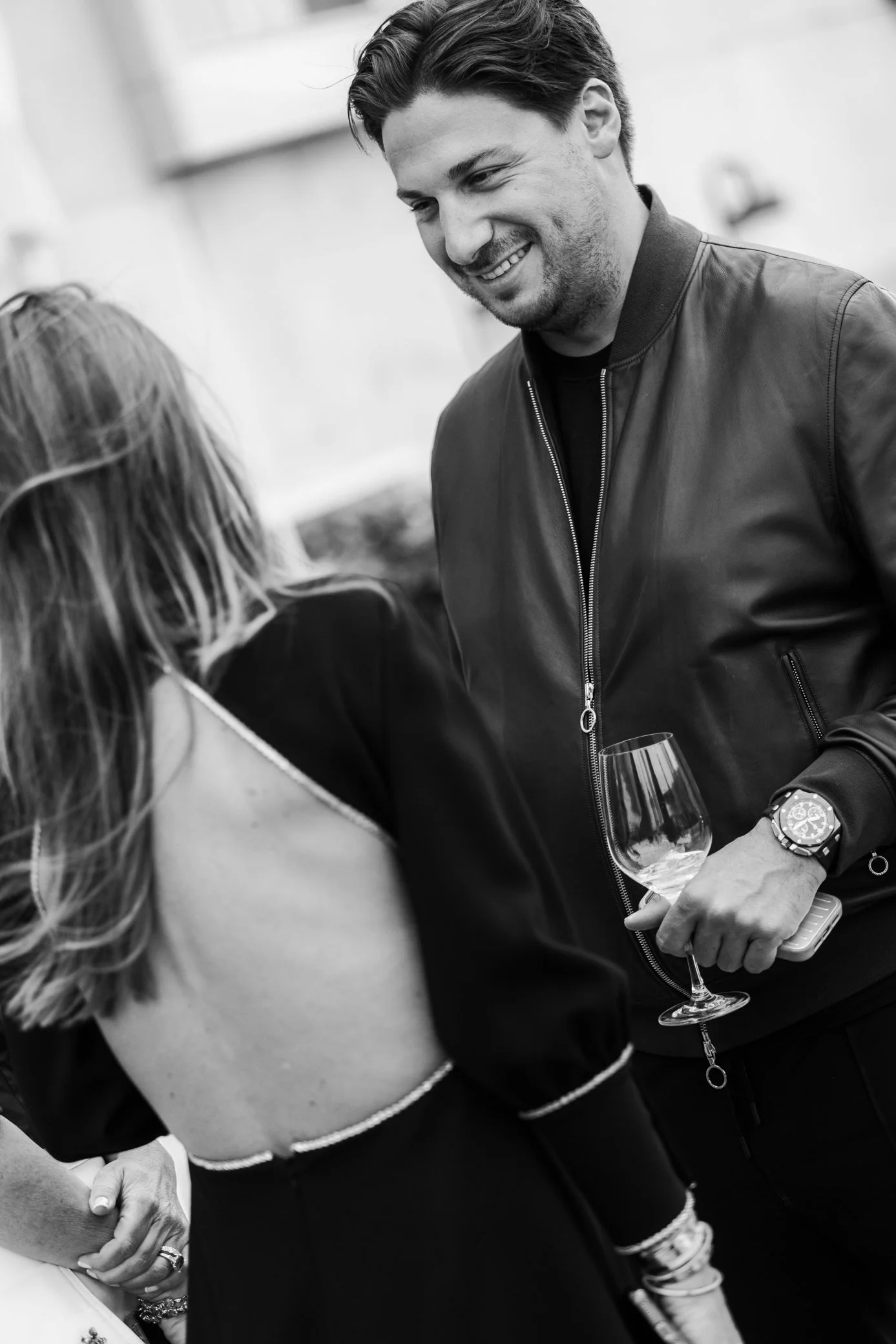Portrait en noir et blanc d’un homme souriant en blouson en cuir, tenant un verre de vin lors d’un événement.