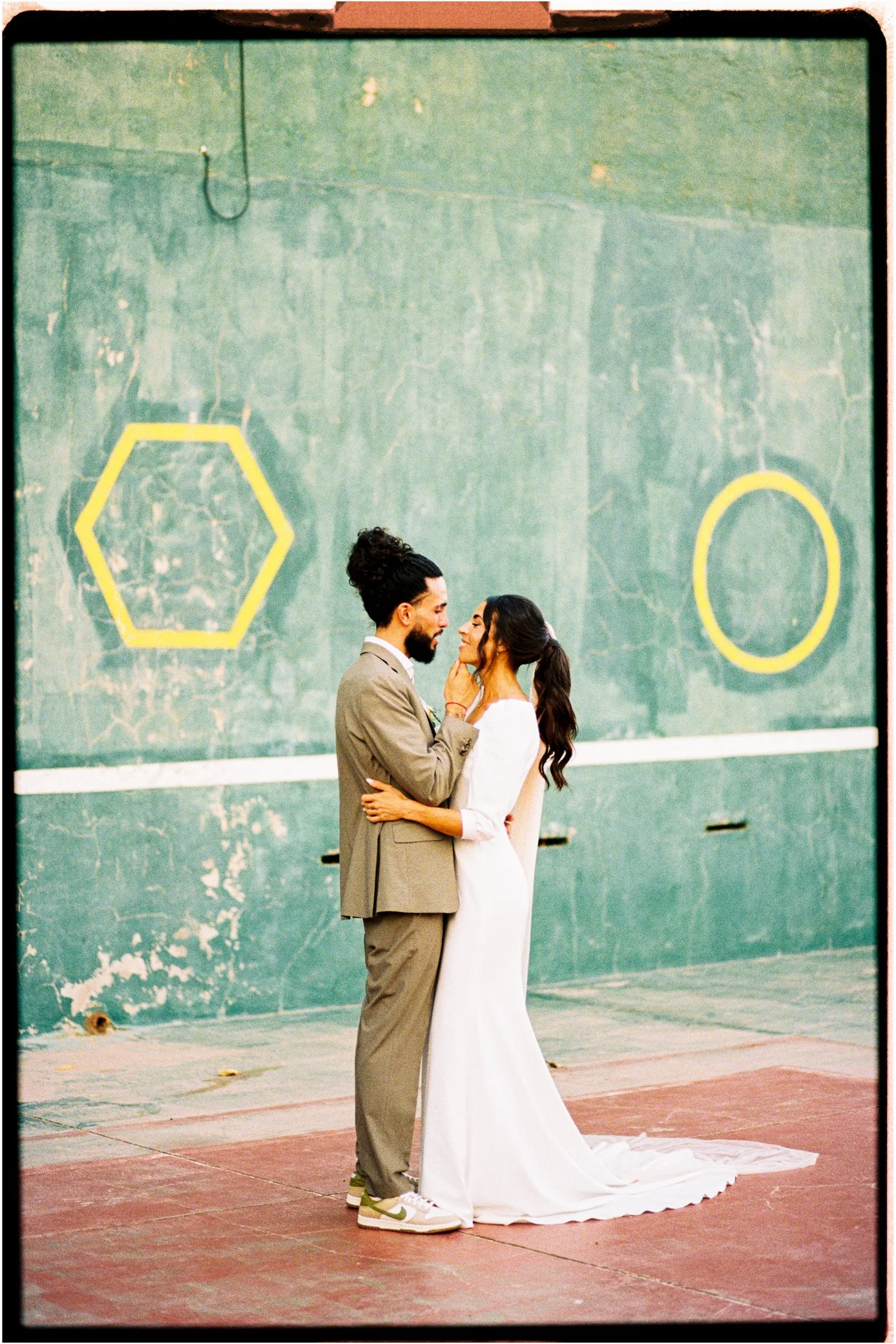 Couple posant tendrement devant un mur vert, l’homme en costume beige et la femme en robe blanche à traîne.