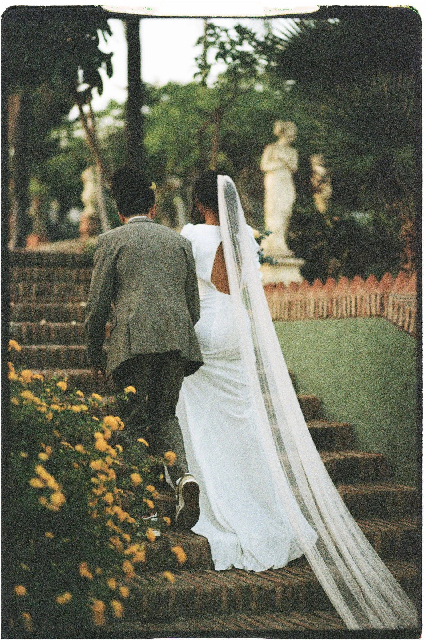 Couple de mariés marchant dans un jardin fleuri, vue de dos, avec la longue traîne de la robe blanche.
