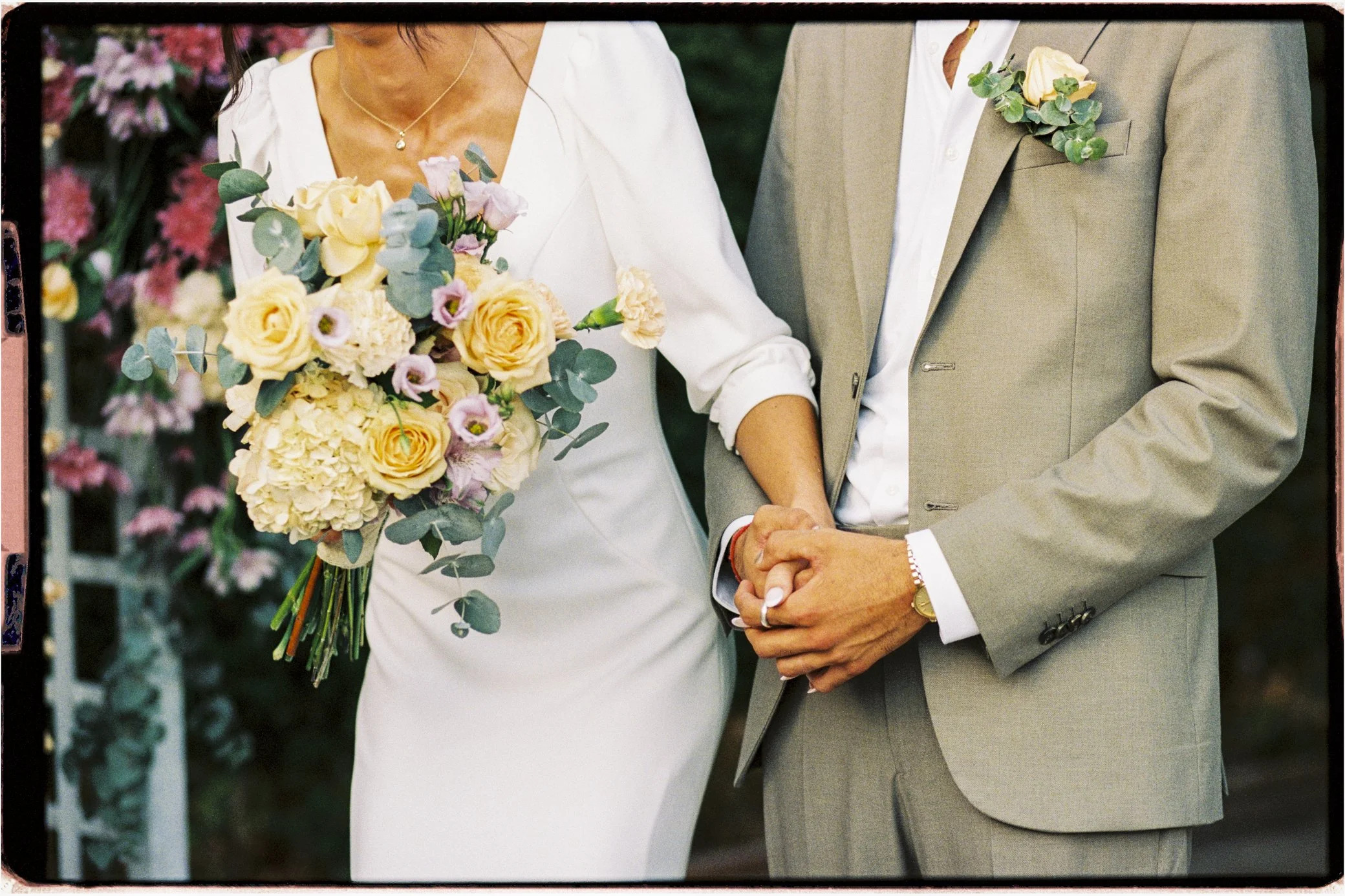 Mariés se tenant la main, la mariée portant un bouquet de fleurs pastel et le marié un costume beige fleuri.