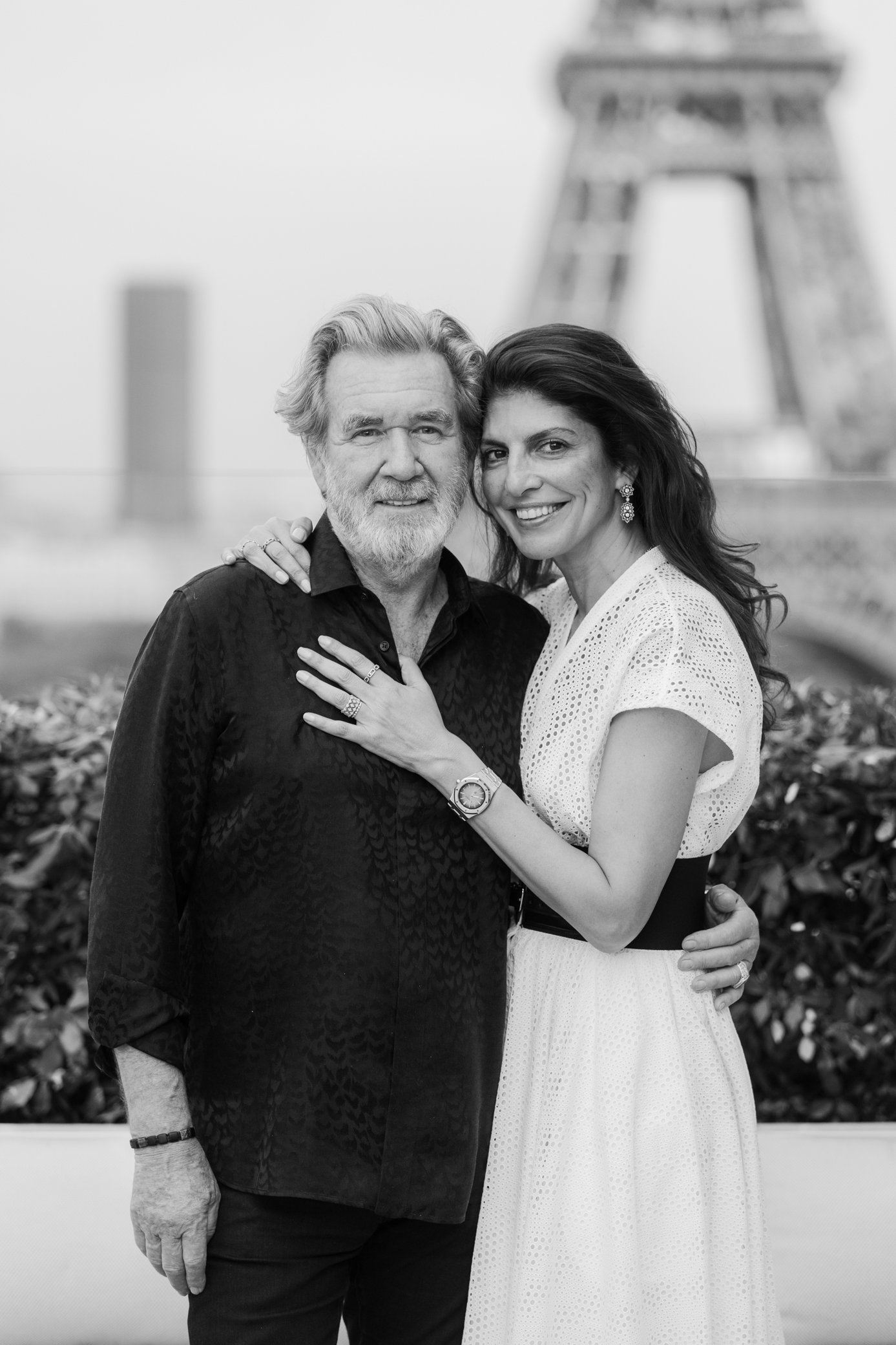 Portrait en noir et blanc d’un couple souriant devant un bâtiment, l’homme portant une chemise sombre et la femme une robe blanche.