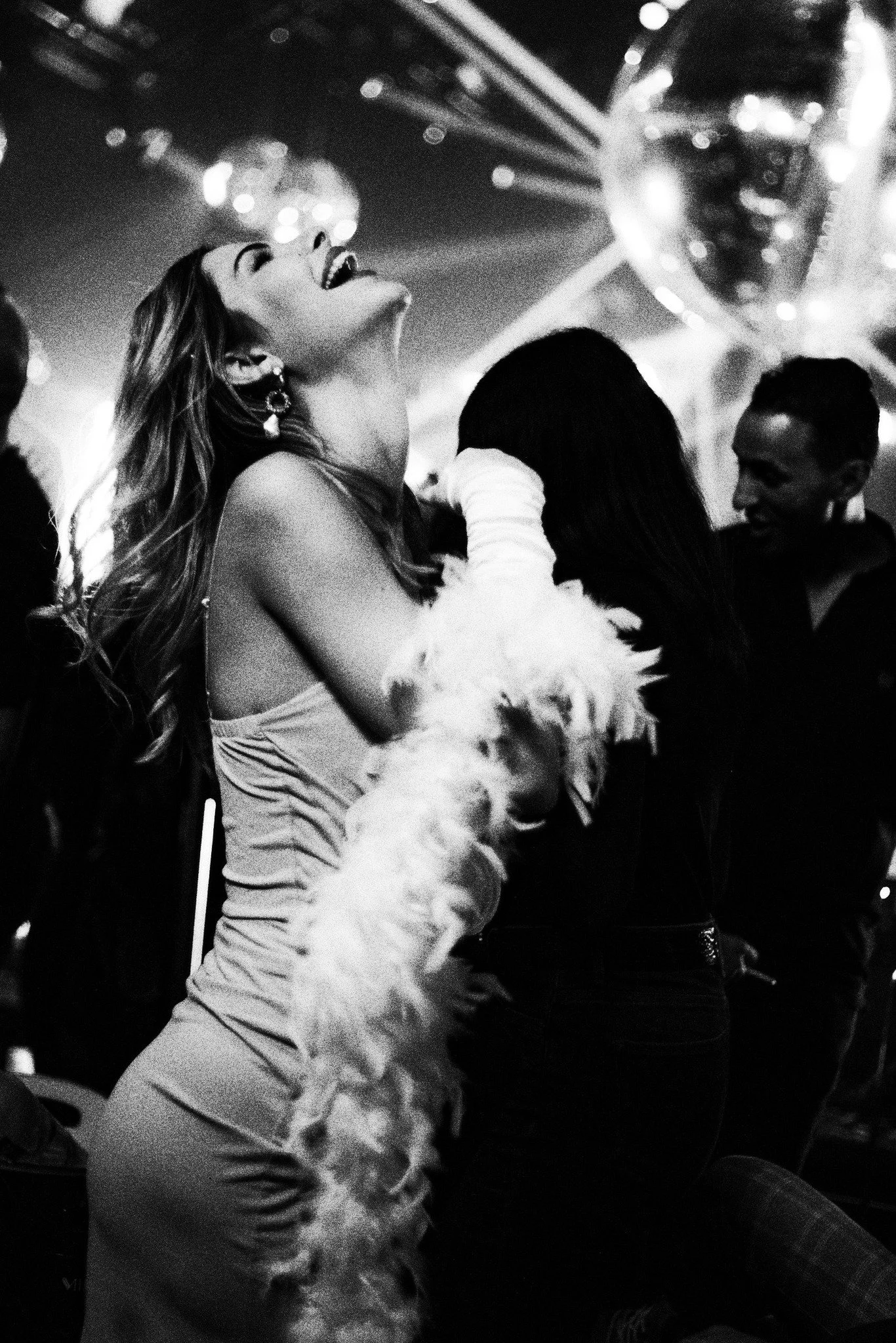 Femme dansant et riant lors d’une soirée festive en noir et blanc, entourée de convives et de lumières scintillantes.