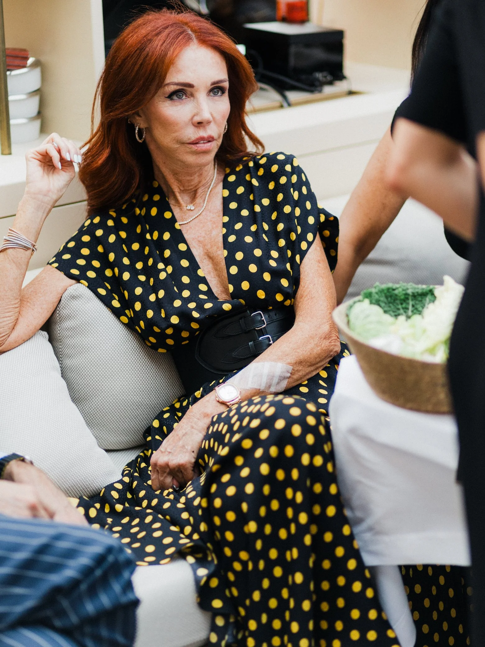 Femme rousse assise sur un canapé, vêtue d’une robe noire à pois jaunes, discutant avec une personne dans un intérieur.