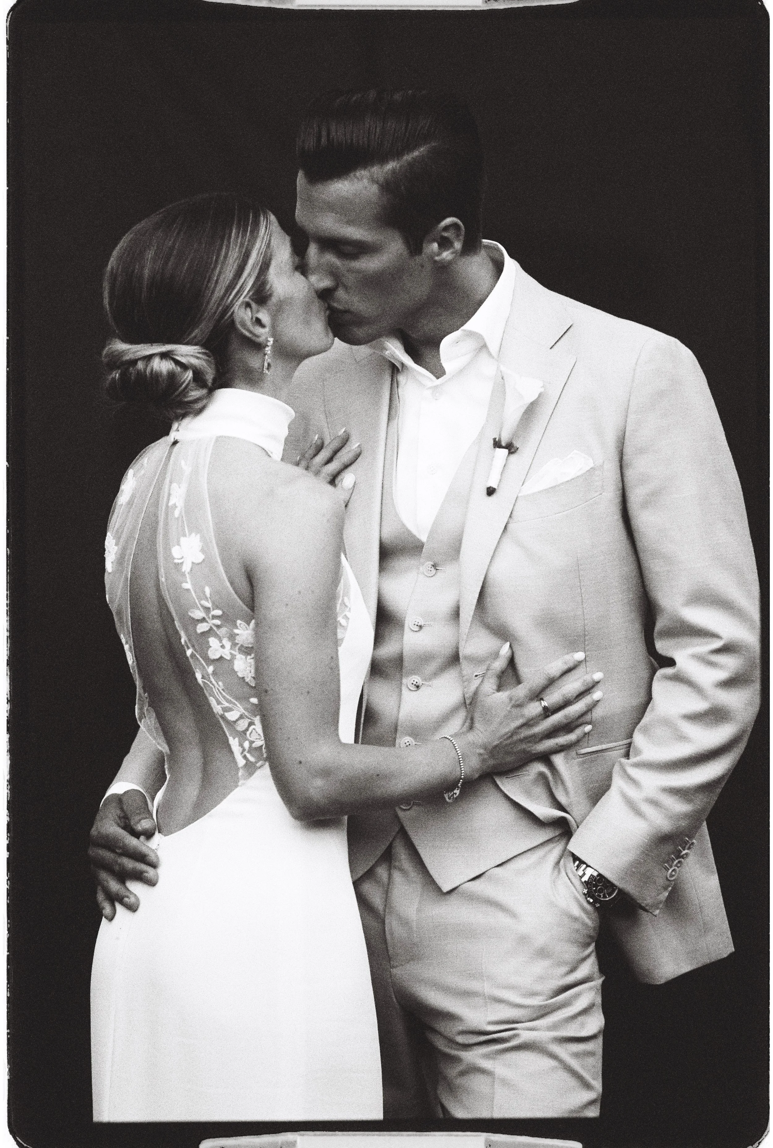 Photo en noir et blanc d’un couple de jeunes mariés s’embrassant, la mariée portant un voile brodé et le marié un costume clair.