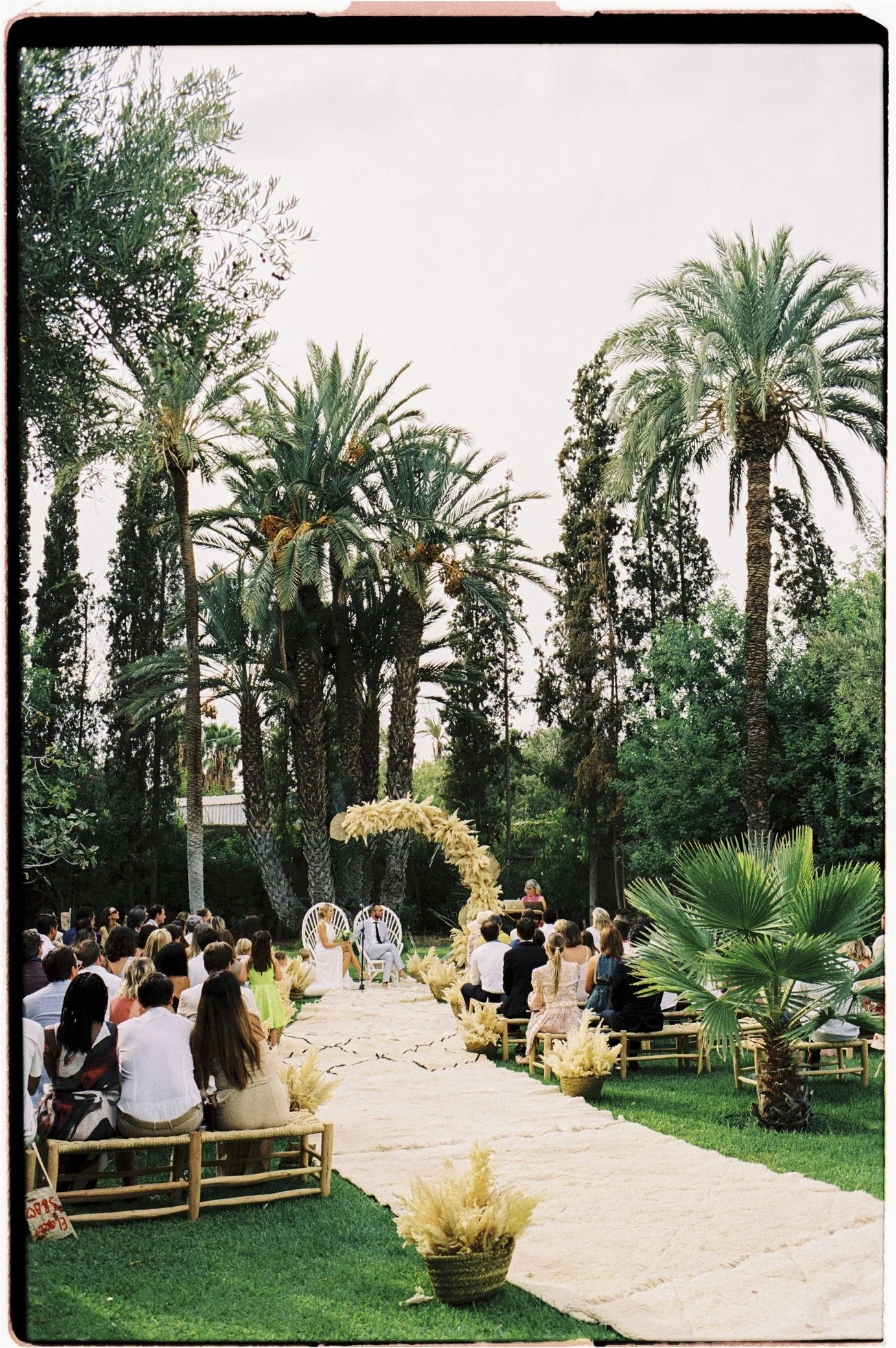 Cérémonie de mariage en plein air dans un jardin luxuriant bordé de palmiers, avec des invités assis face aux mariés.