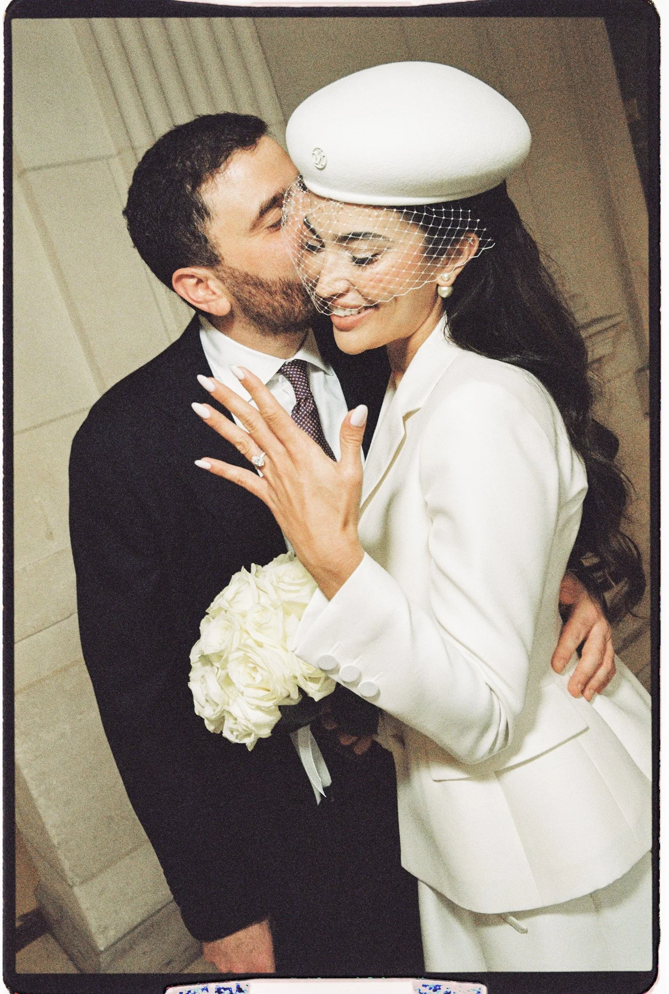 Couple de mariés enlacés, la mariée en tailleur blanc et chapeau voilé tenant un bouquet de roses blanches.