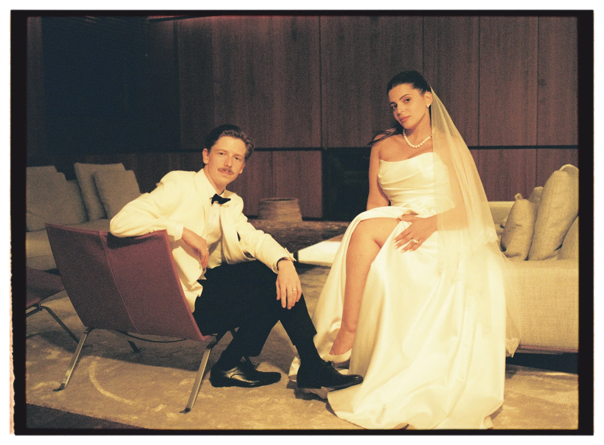 Un couple pose dans un salon : un homme en costume blanc assis dans un fauteuil et une femme en robe de mariée avec voile assise sur un canapé.