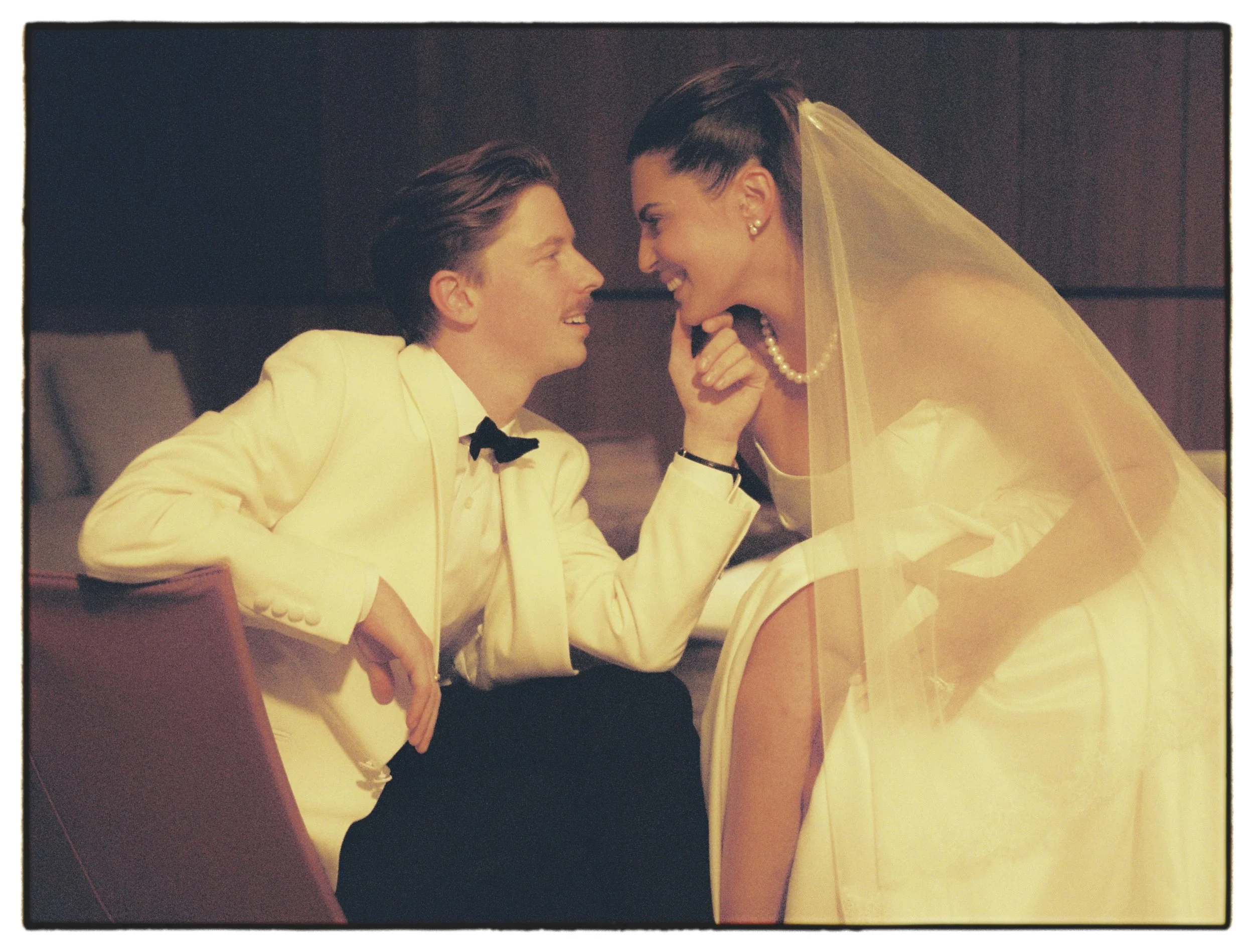 Jeune couple de mariés souriant et se regardant tendrement, l’homme en costume blanc et nœud papillon, la femme en robe blanche et voile.