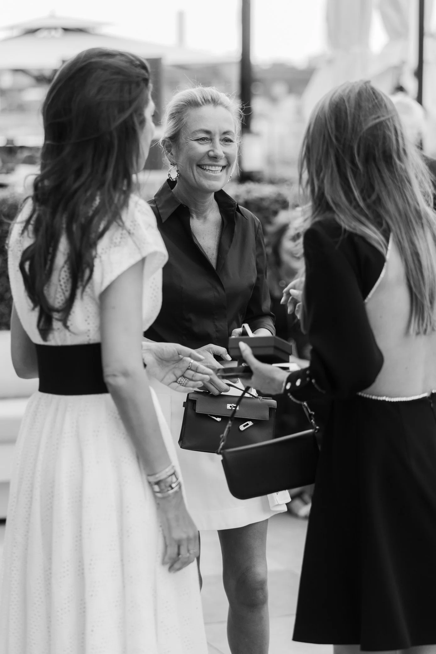 Trois femmes discutent lors d’un événement élégant, tandis que la femme au centre sourit, en noir et blanc.