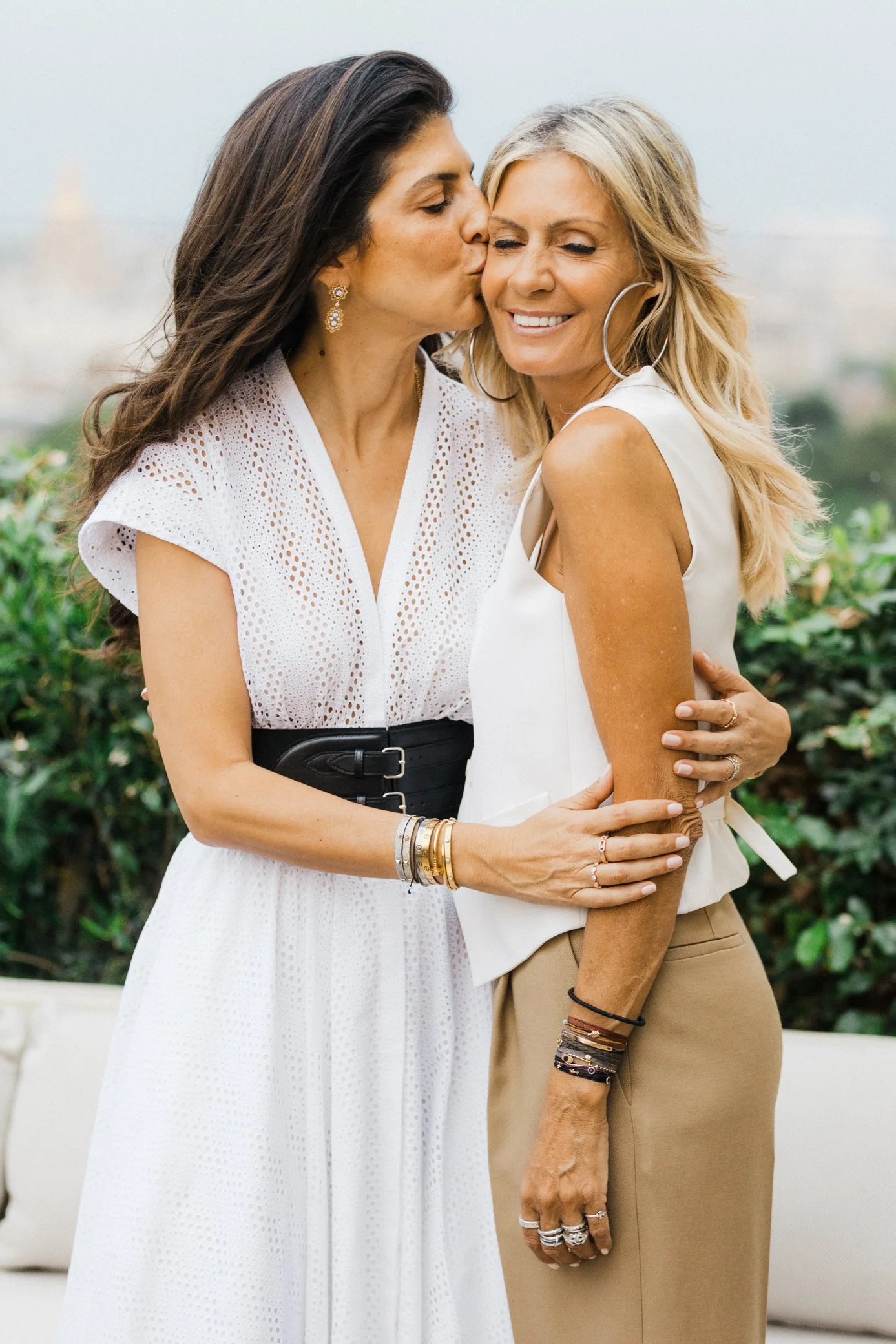 Deux femmes souriantes en tenue blanche, enlacées dans un jardin, dont l’une embrasse affectueusement l’autre sur la joue.