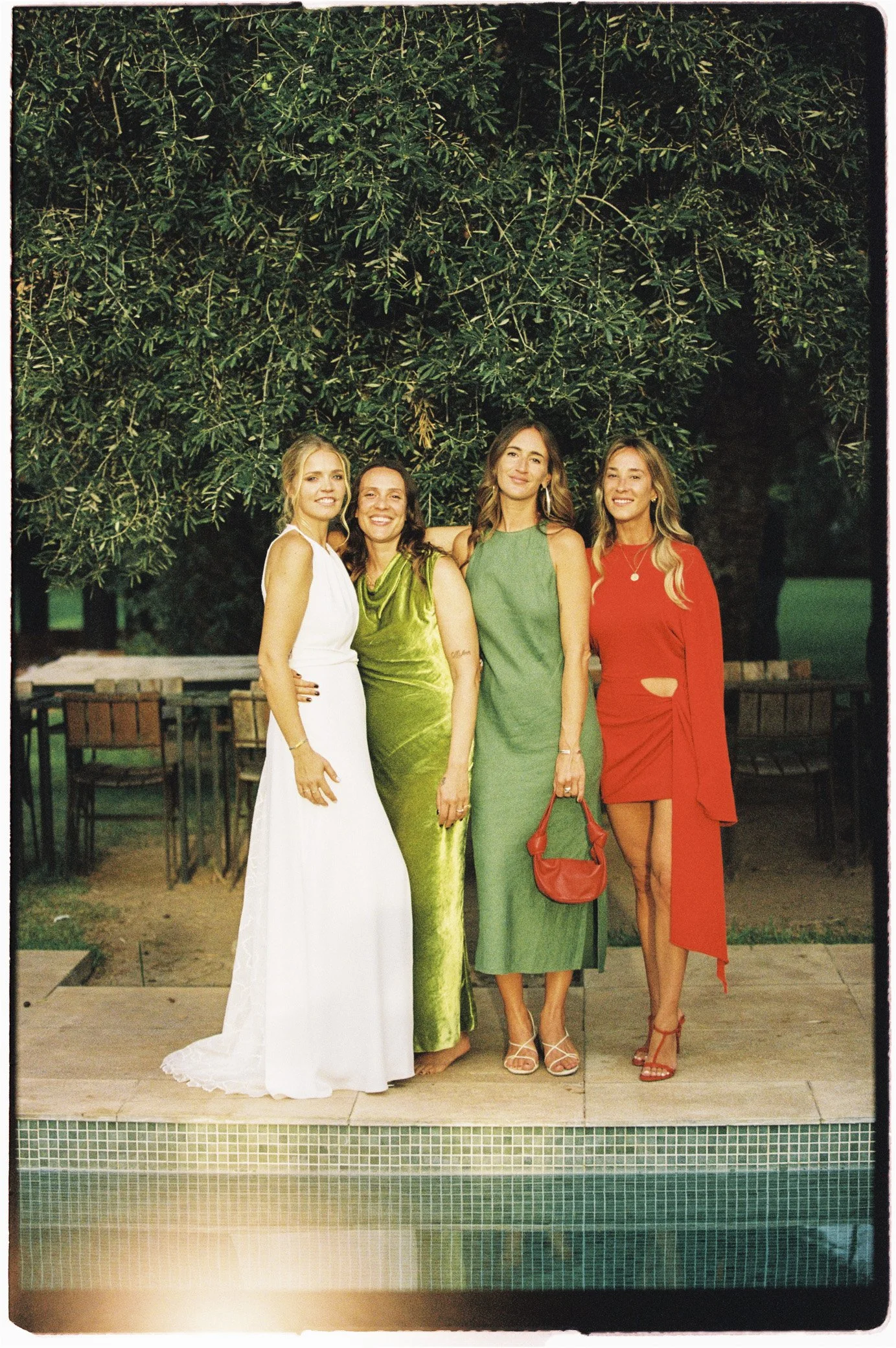 Quatre femmes posant au bord d’une piscine, vêtues de robes élégantes blanches, vertes et rouges, dans un jardin arboré.