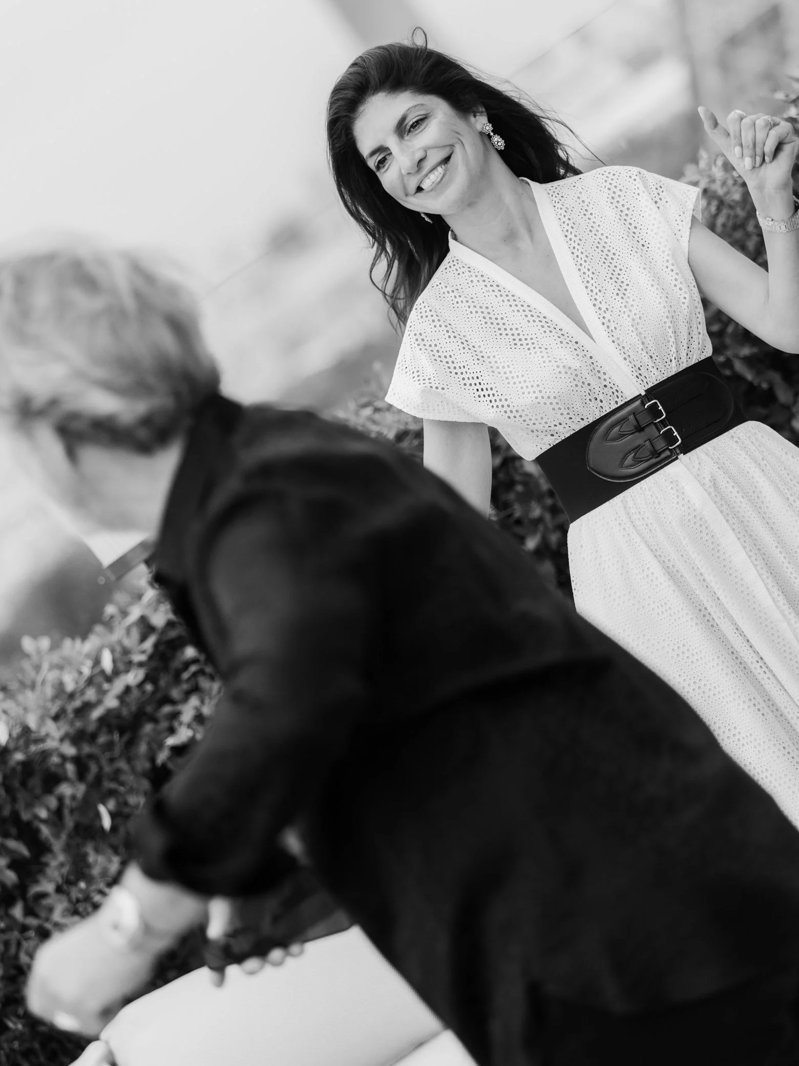 Portrait en noir et blanc d’une femme souriante en robe blanche ajourée et large ceinture, entourée de personnes floues en extérieur.