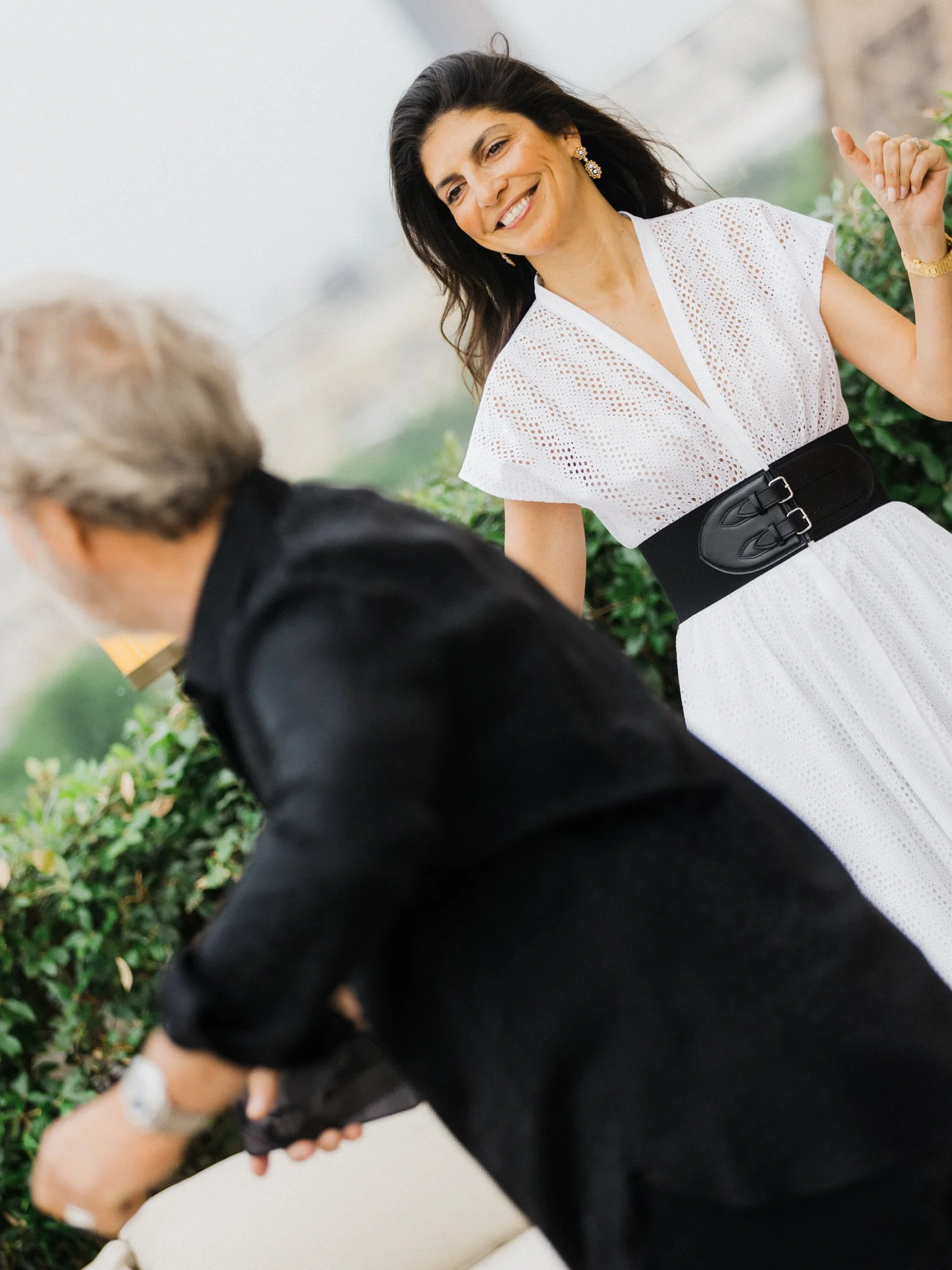 Femme souriante en robe blanche ajourée et ceinture noire, debout à côté d’une personne vêtue de noir dans un jardin.