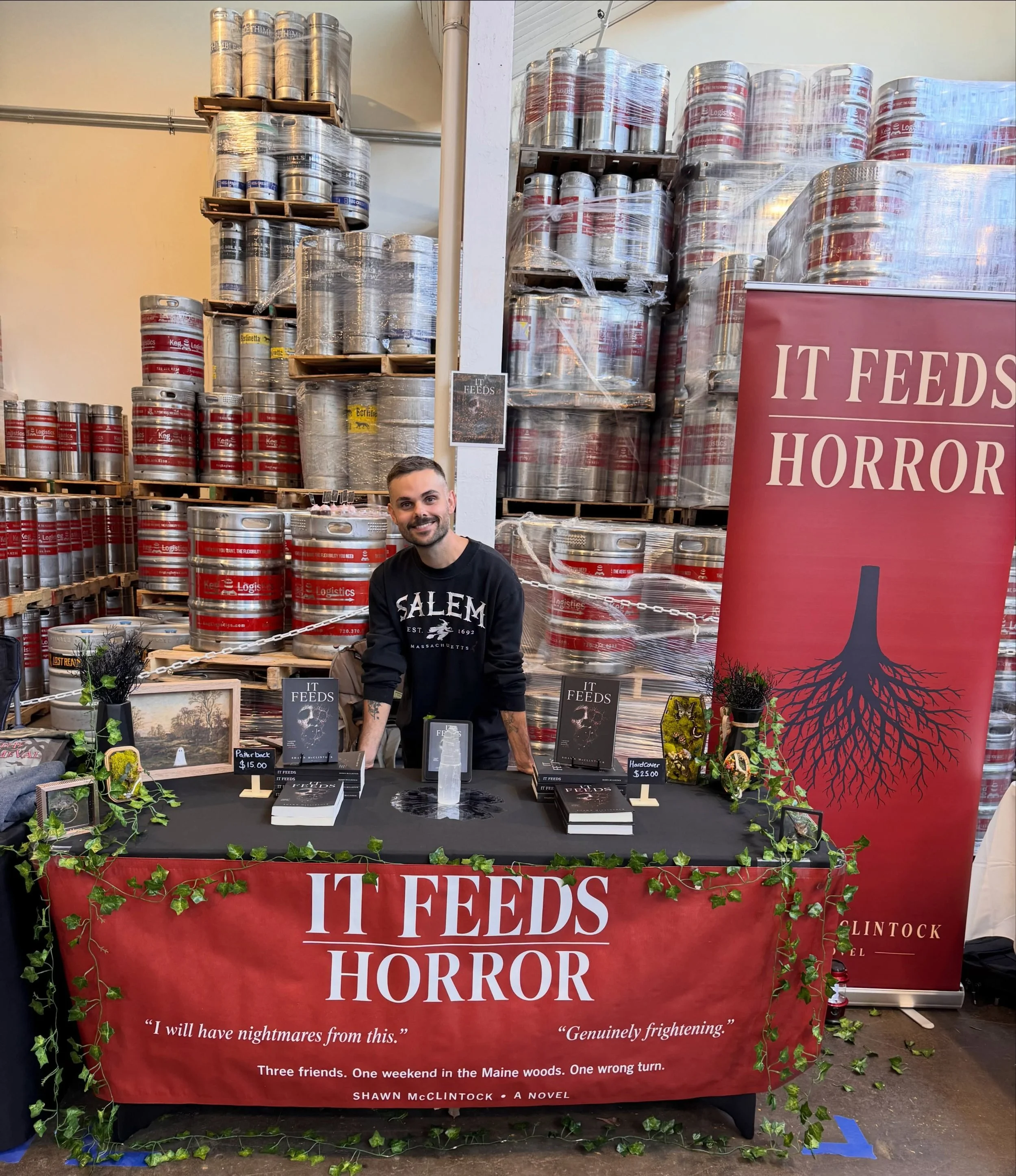 Author Shawn McClintock stands at a book signing table for his horror novel “It Feeds,” surrounded by stacked beer kegs and promotional displays.