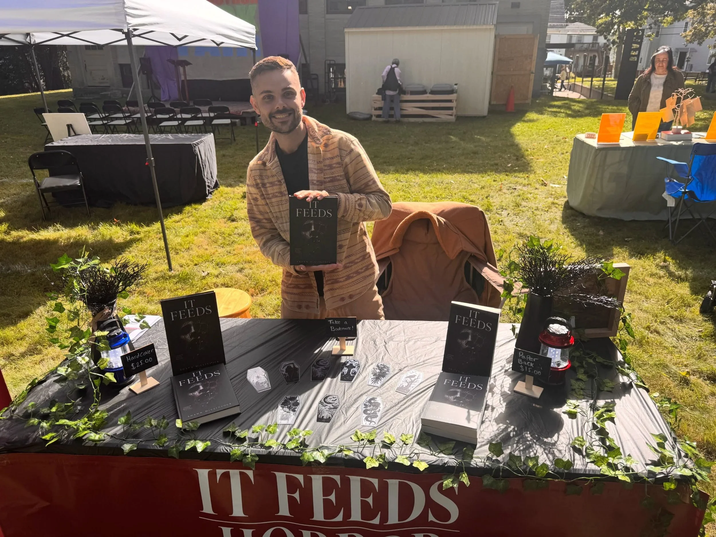 Author displaying a book at an outdoor literary market booth with books, art cards, and “It Feeds” signage.